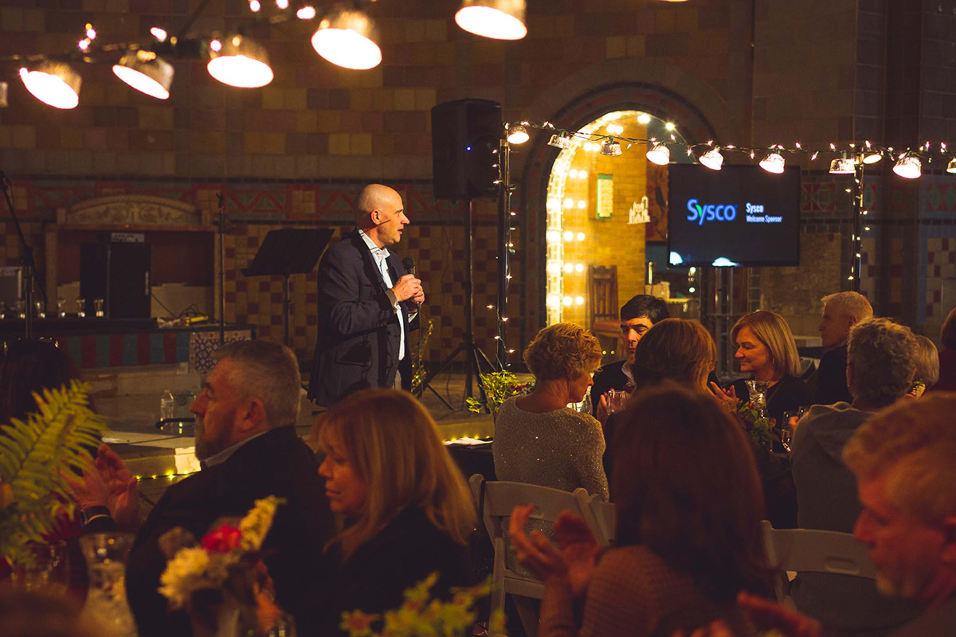 A man stands speaking at a podium in a decorated event space with guests seated at tables.