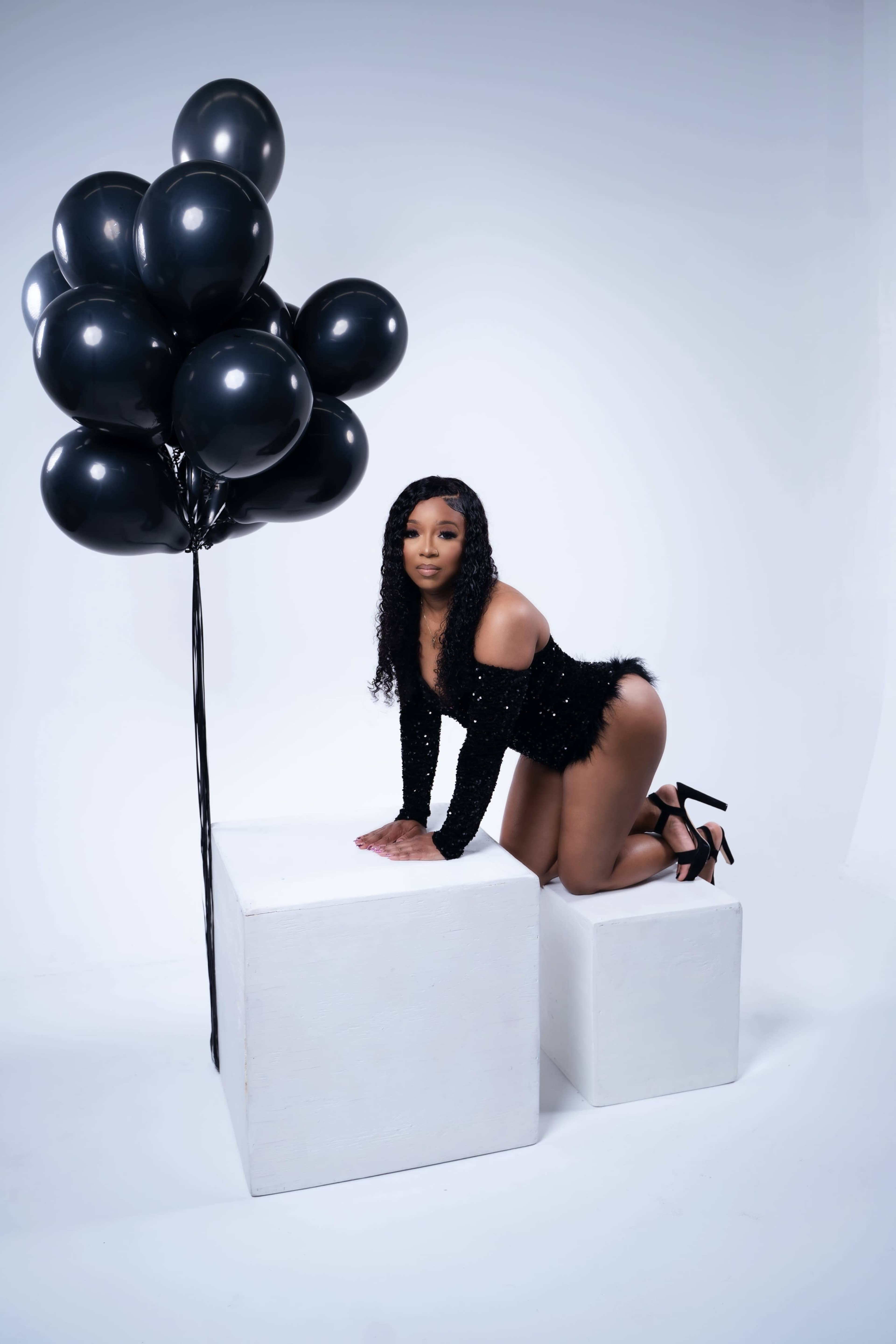 A woman in a black outfit kneels on two white cubes beside a cluster of black balloons.