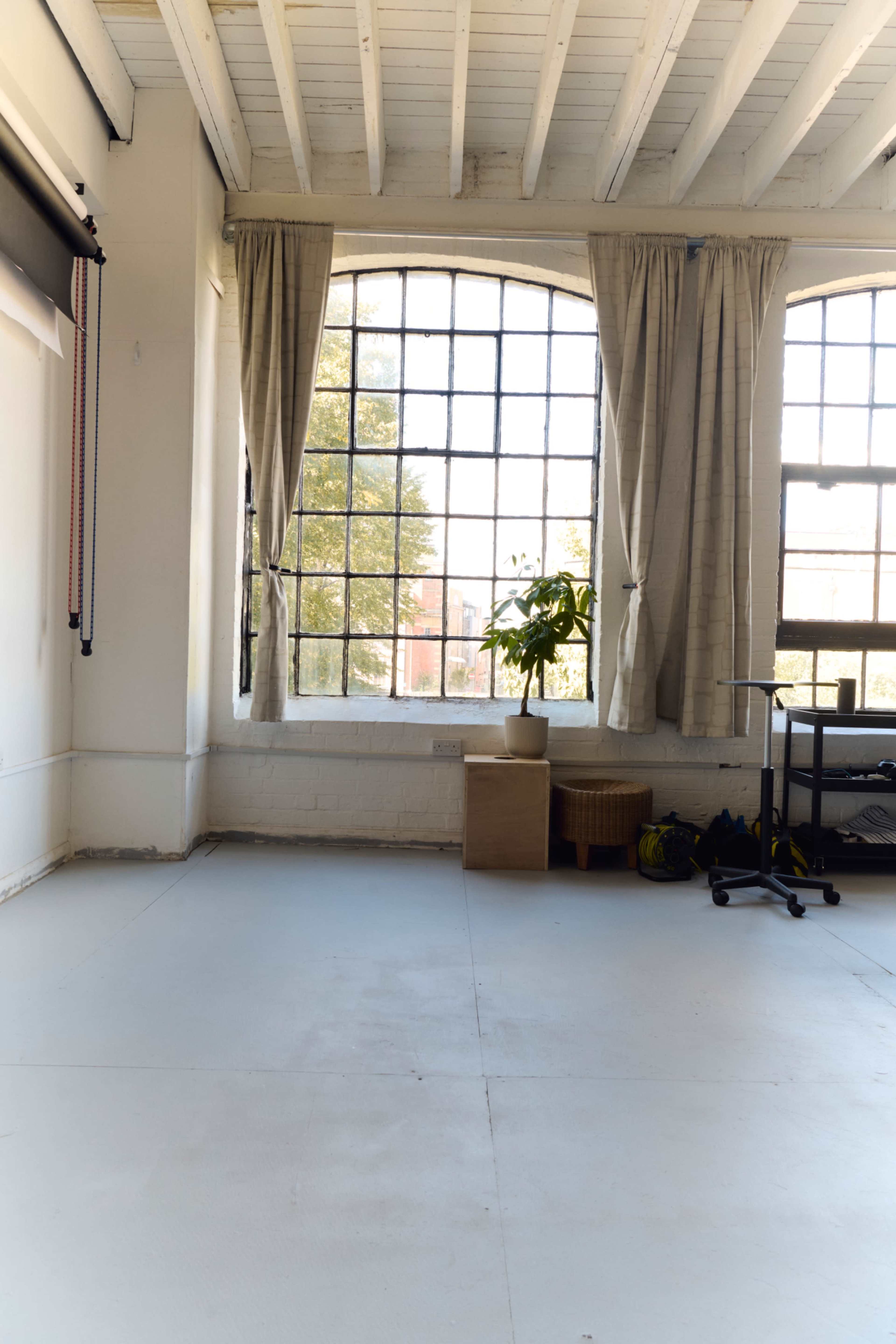 The image shows a bright, spacious room with large windows, light-colored walls, and minimal furnishings, including a potted plant and a small storage box.