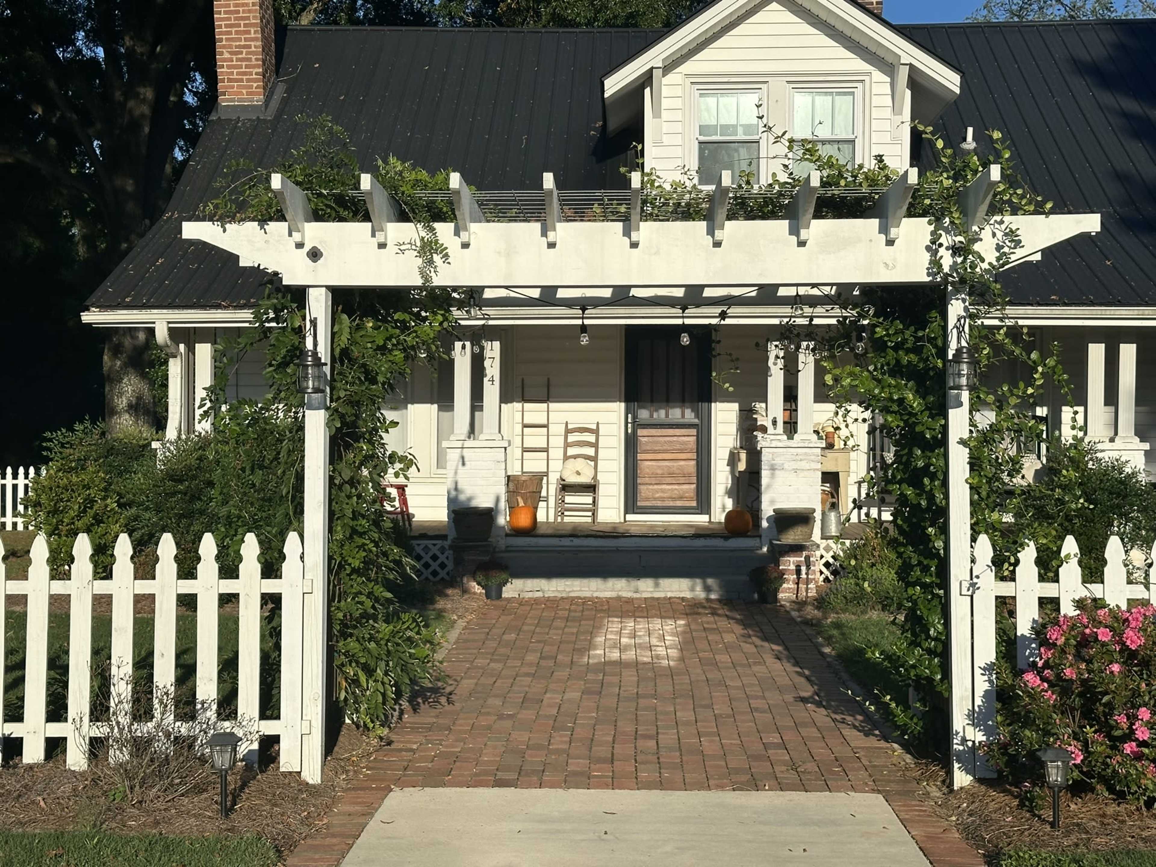 The image shows a white house with a covered porch, surrounded by a picket fence and landscaped with plants and flowers.