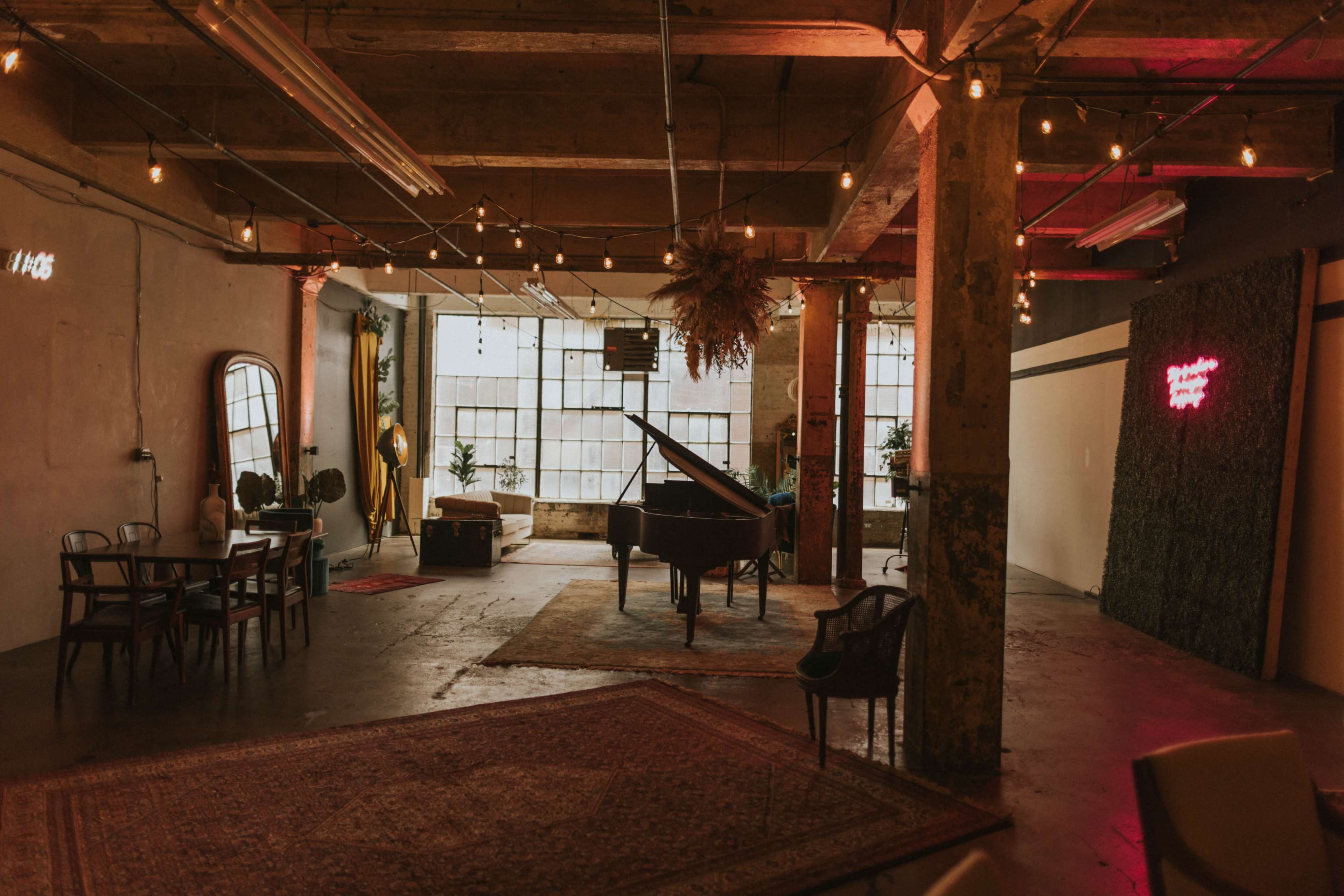 The image shows an industrial-style space featuring a grand piano, a dining table, and decorative lighting with plants and textured walls.