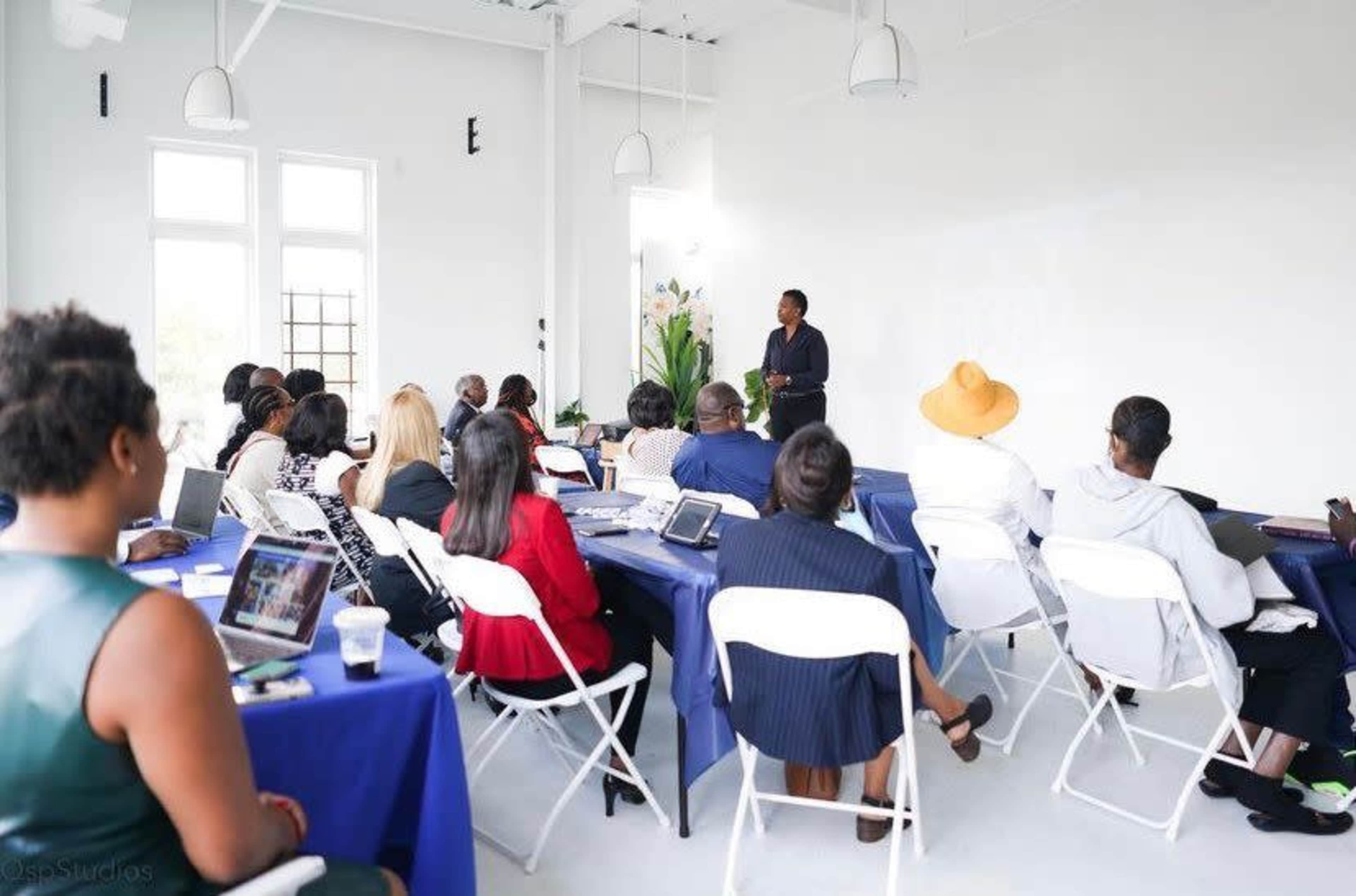 A speaker presents to an audience seated at tables in a well-lit meeting room.