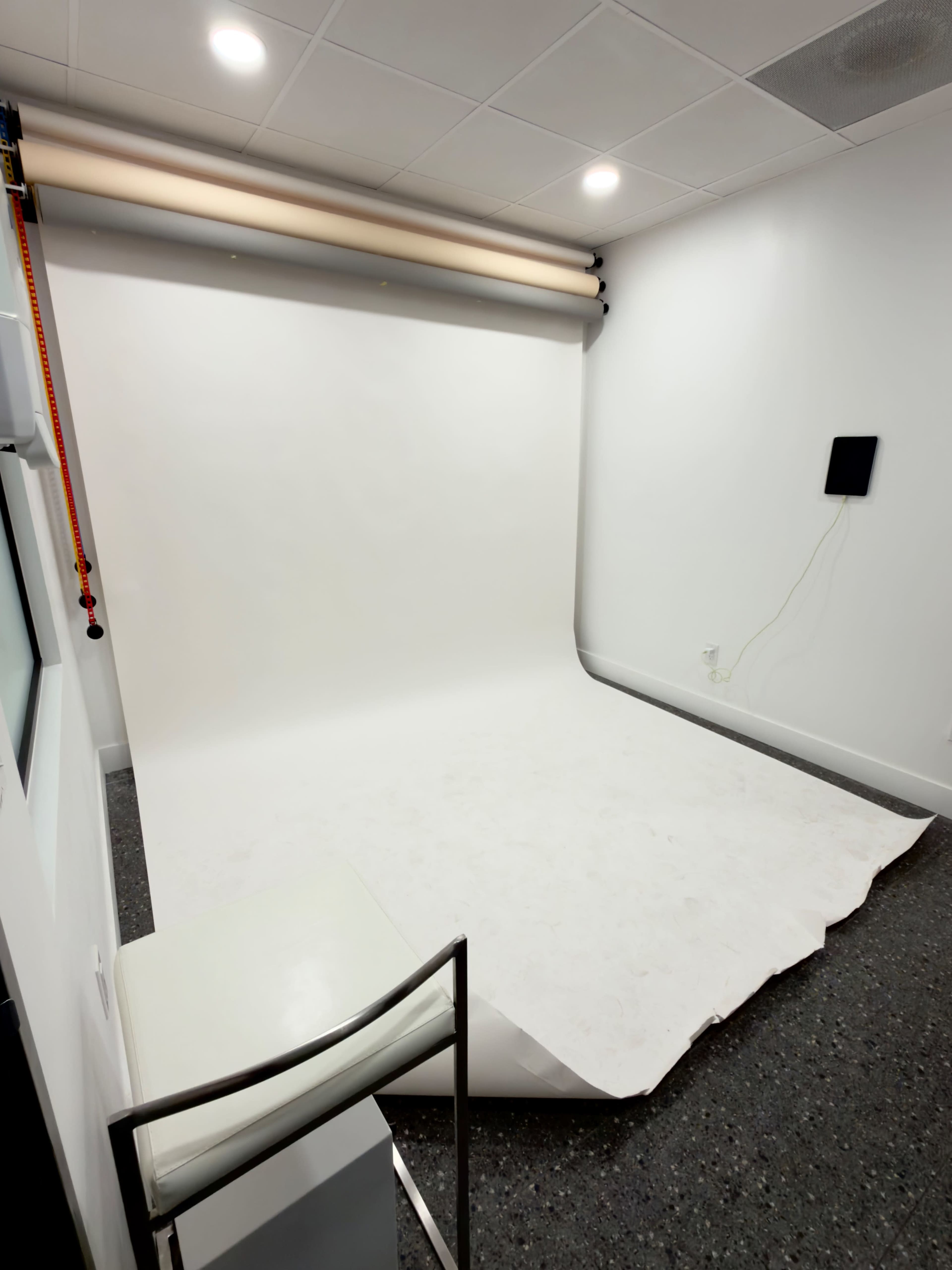 The image shows a photography studio with a white backdrop, a chair, and a small wall-mounted monitor.