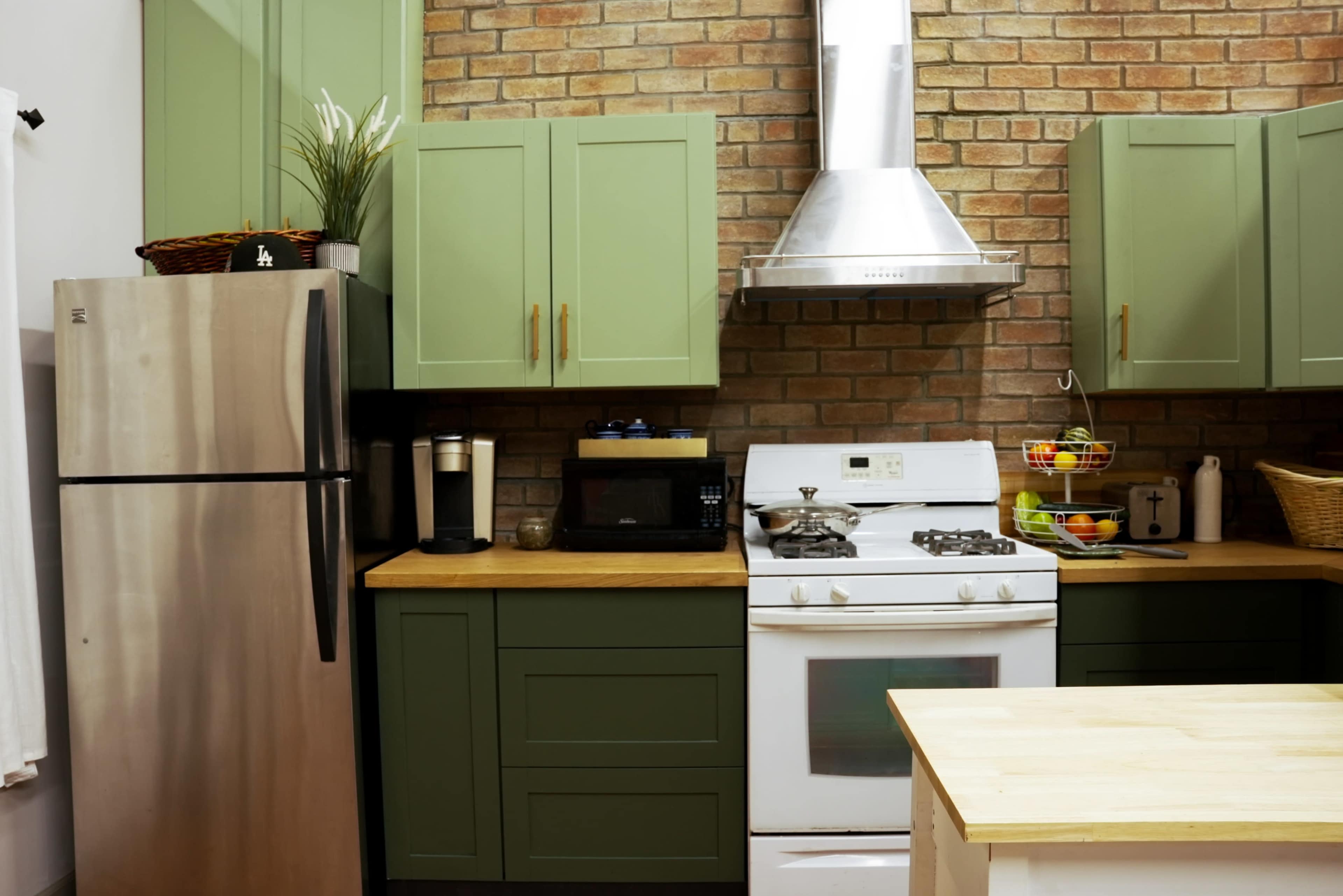 The kitchen features green cabinets, a stainless steel refrigerator, a gas stove, and a brick wall with a silver range hood.