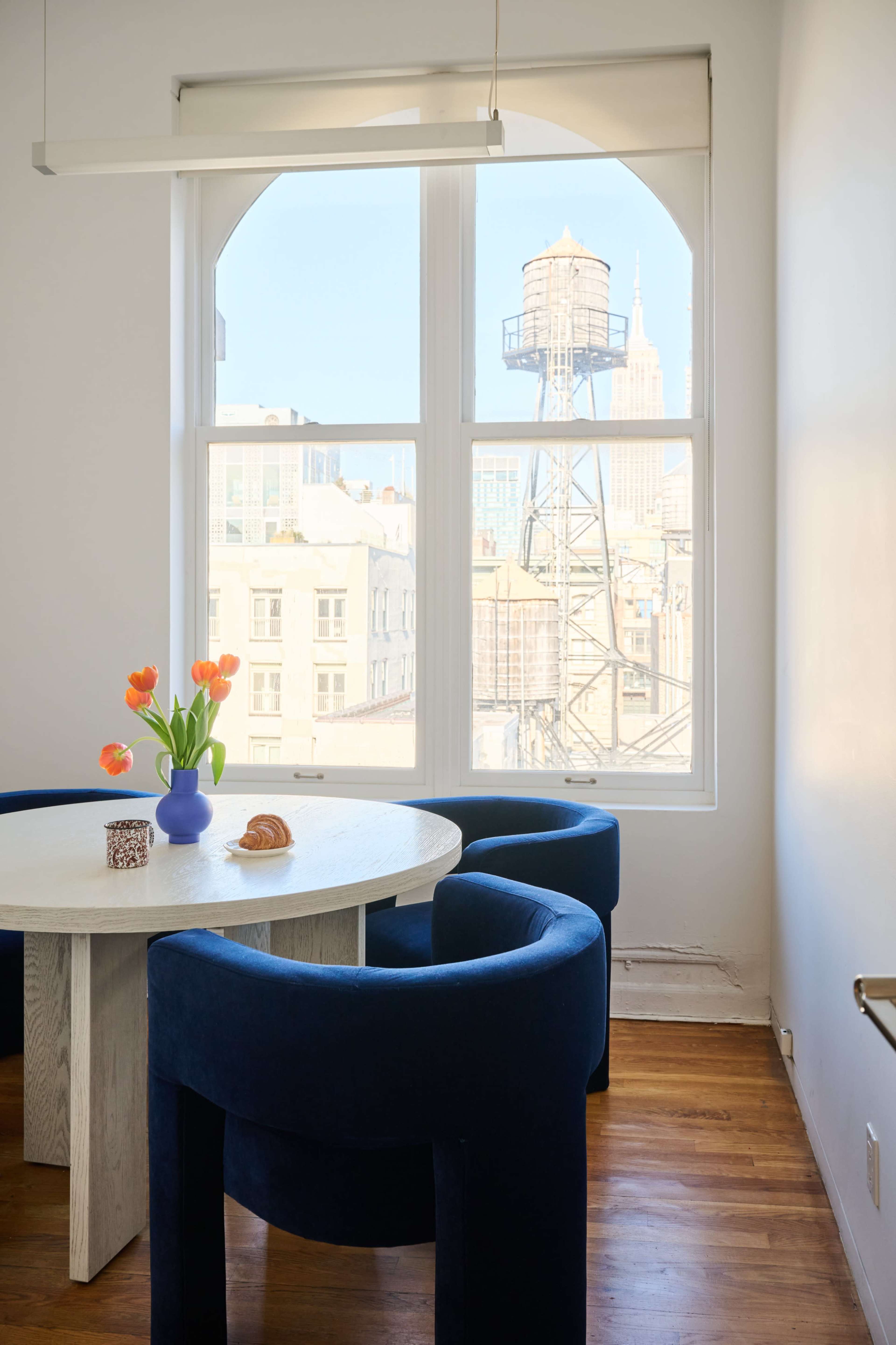 A round table with blue chairs is set in a bright room featuring a view of the Empire State Building and a water tower through large windows.