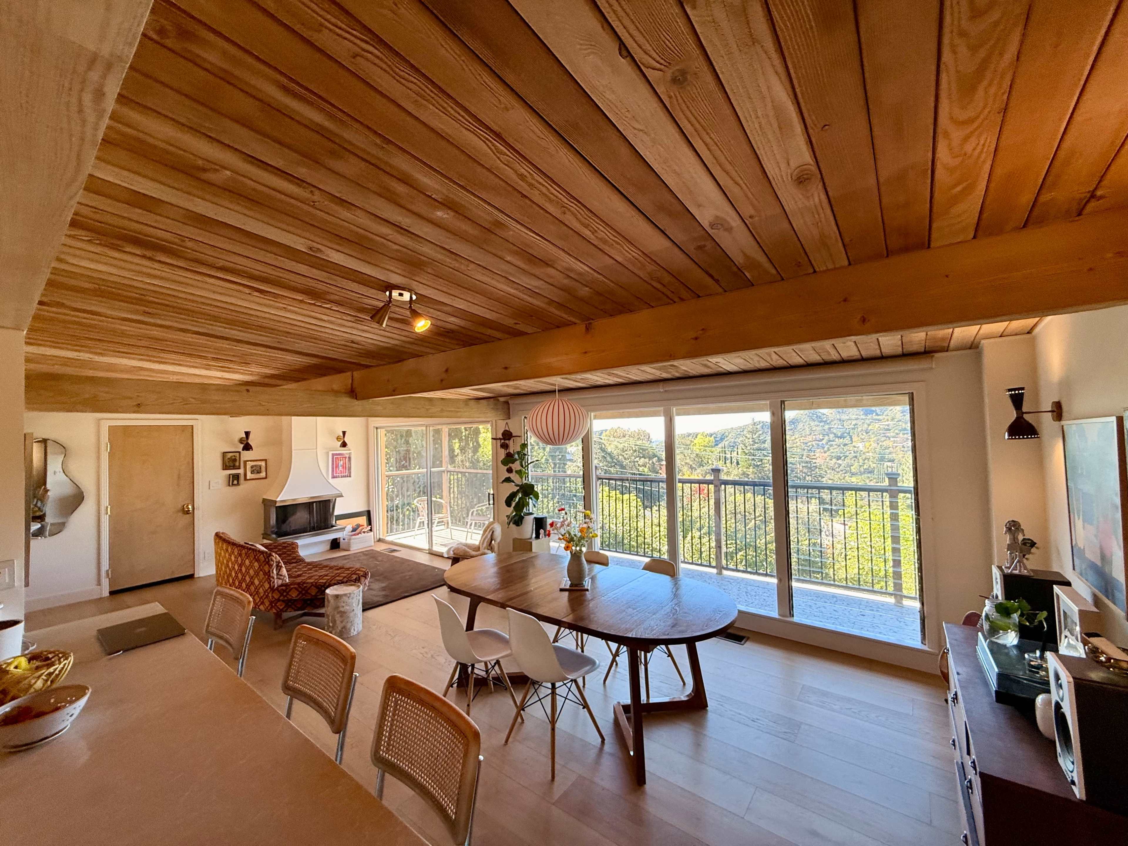 The image depicts a spacious, modern open-concept living area with a wooden ceiling, large windows offering a view of the outdoors, and a dining table surrounded by various chairs.