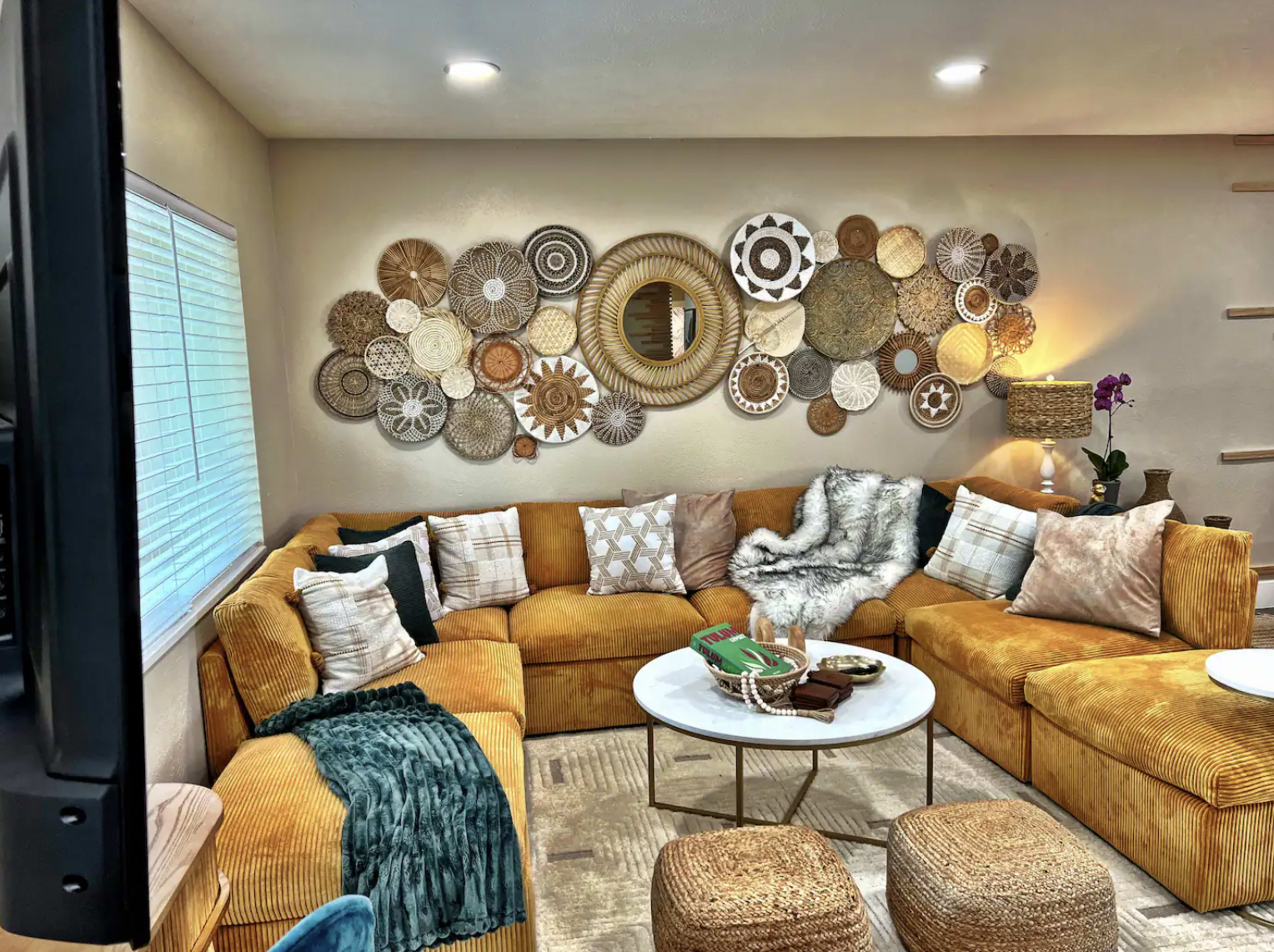 A cozy living room featuring a large, L-shaped, mustard-colored couch adorned with various patterned pillows, a round coffee table, and decorative wall art made of woven materials.