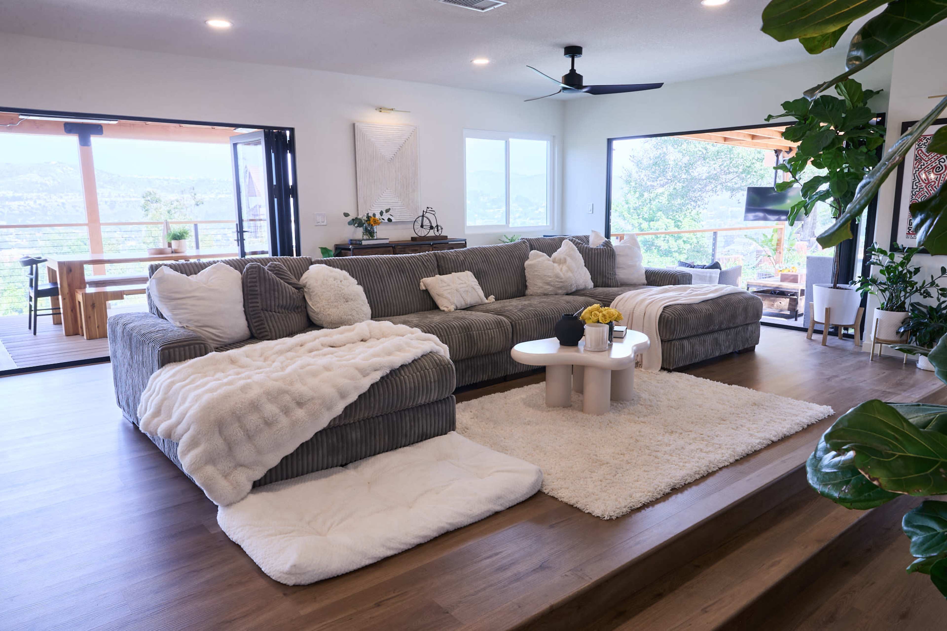 A spacious living room with a large sectional sofa, a round coffee table, and a soft area rug, leading to a deck with a scenic view.