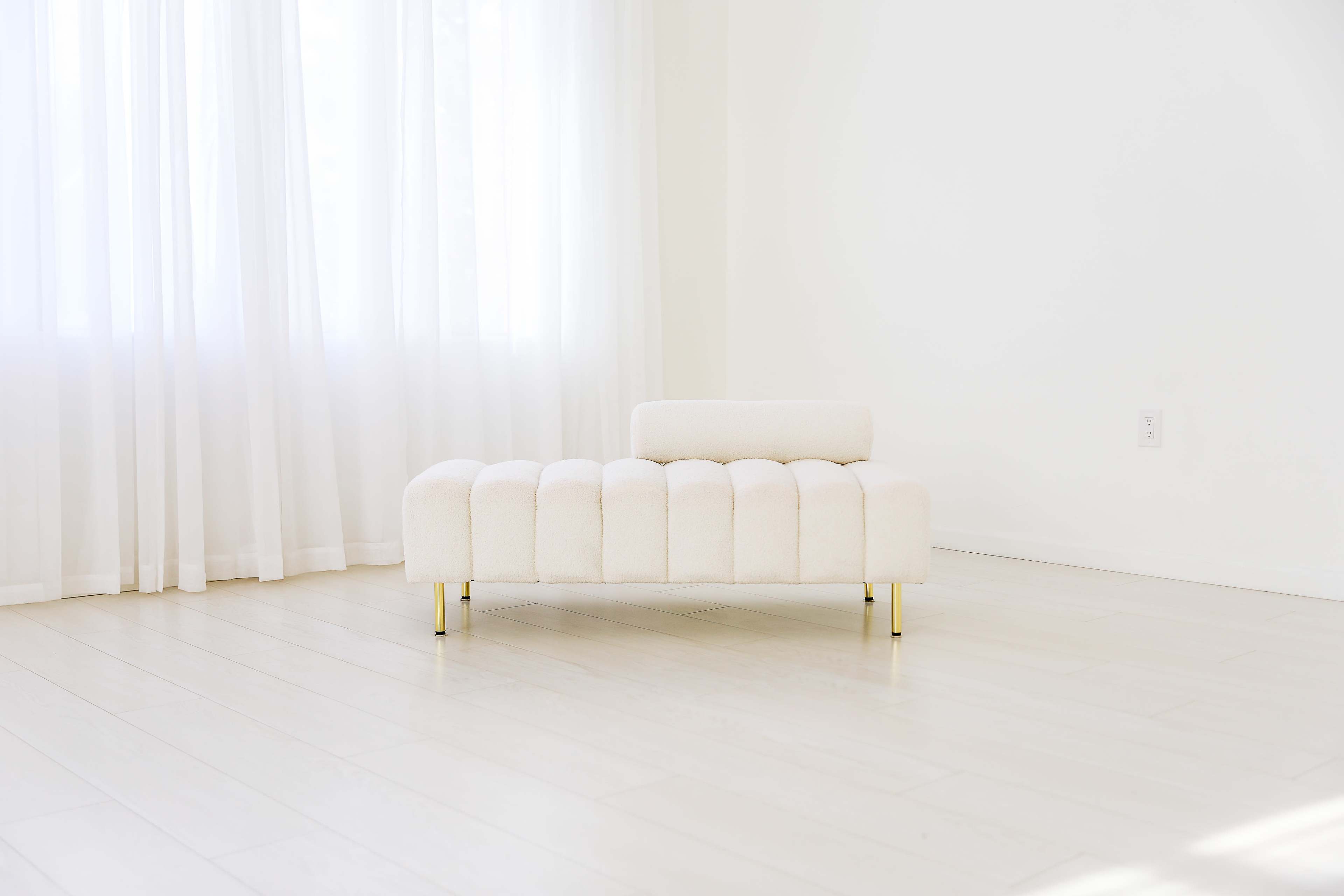 The image shows a light-colored, tufted chaise lounge positioned in a bright, minimally decorated room with large windows.