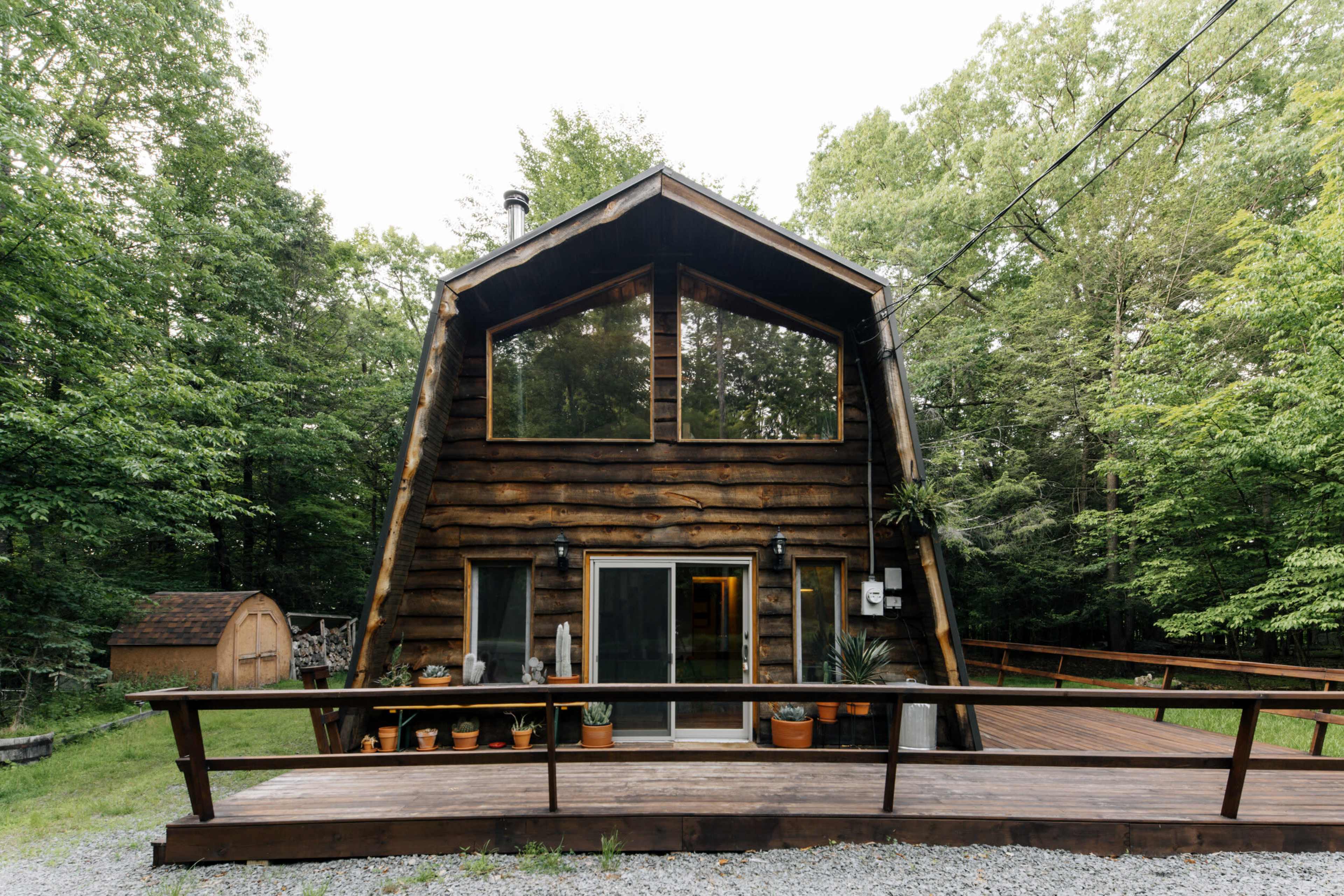 A wooden cabin with a peaked roof is set in a forested area, featuring a front deck and large windows.