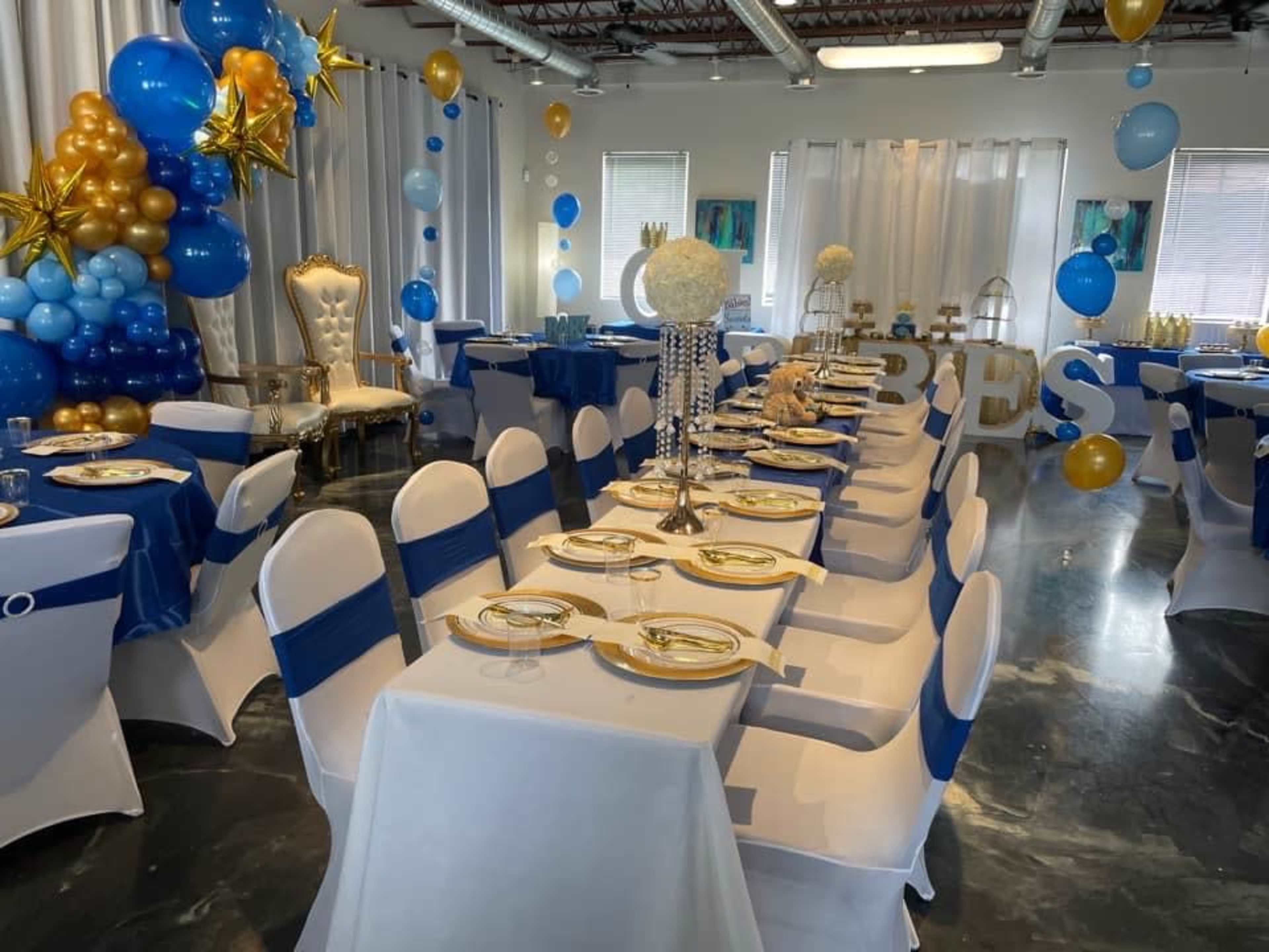A decorated event space with tables set for a celebration, featuring blue and gold balloons, elegant tablecloths, and a royal throne chair.