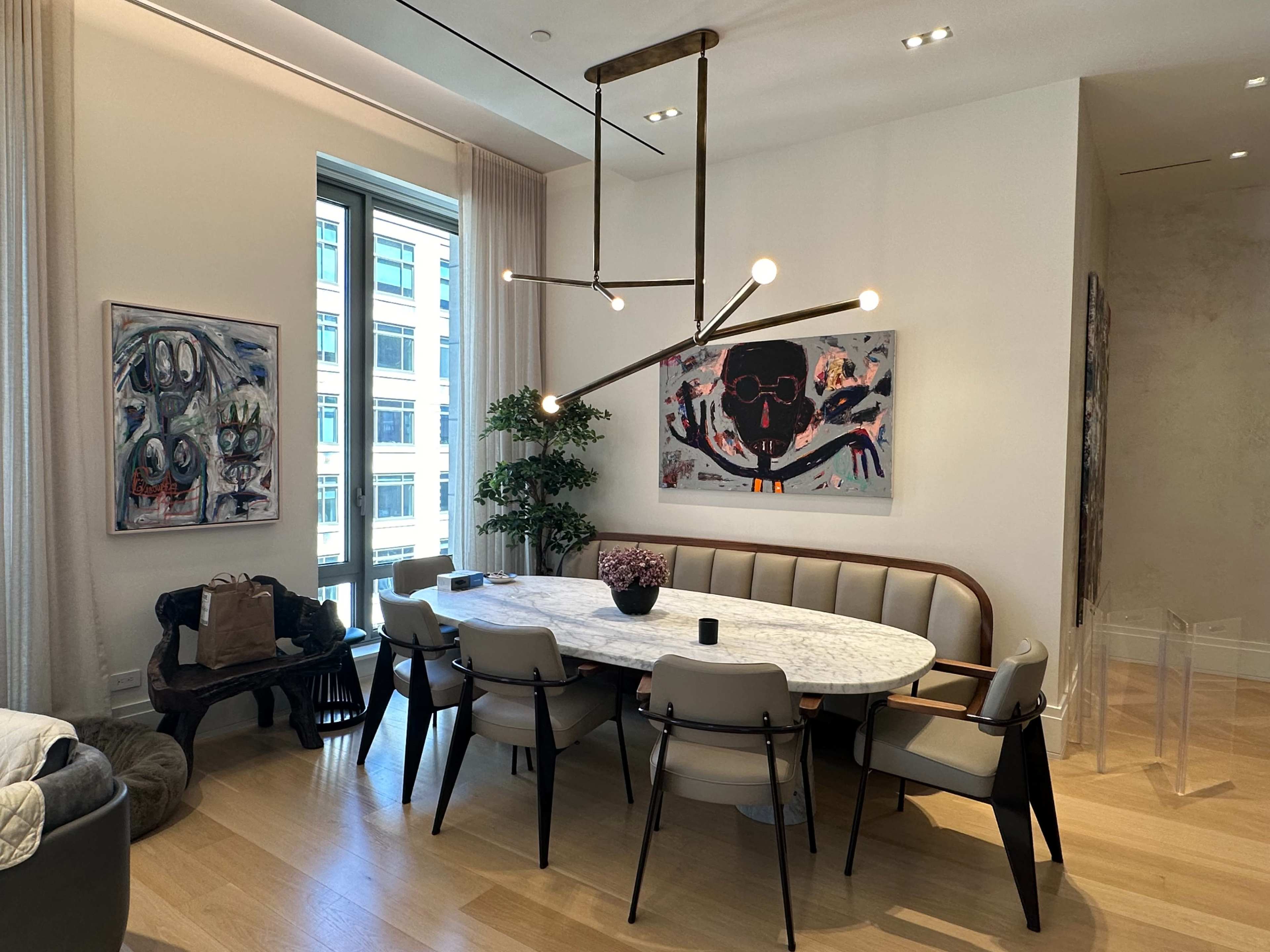 A dining room features a round marble table surrounded by chairs, with abstract artwork on the walls and large windows letting in natural light.