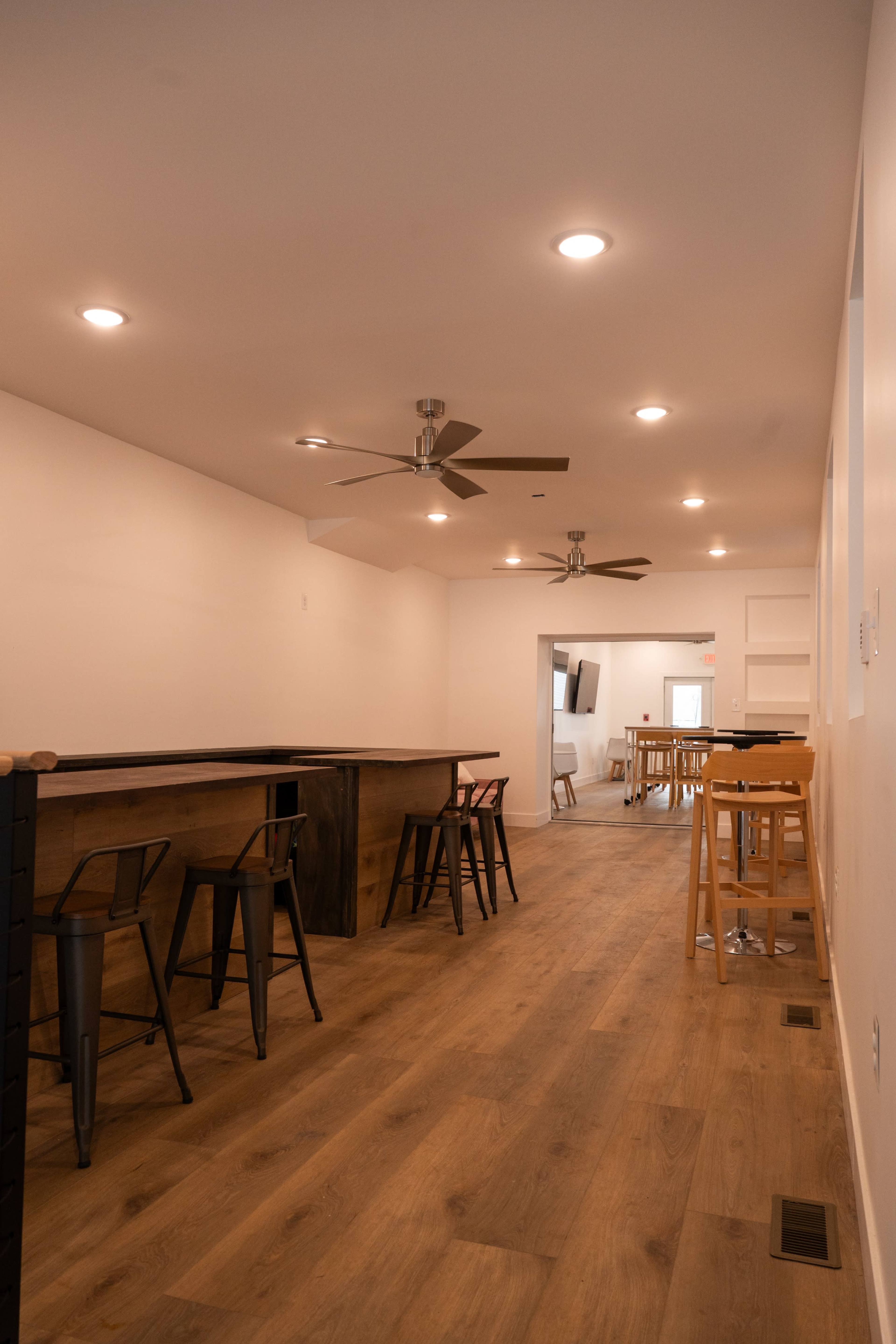 The image shows a long, modern interior space featuring wooden flooring, a bar area with high stools, and ceiling fans.