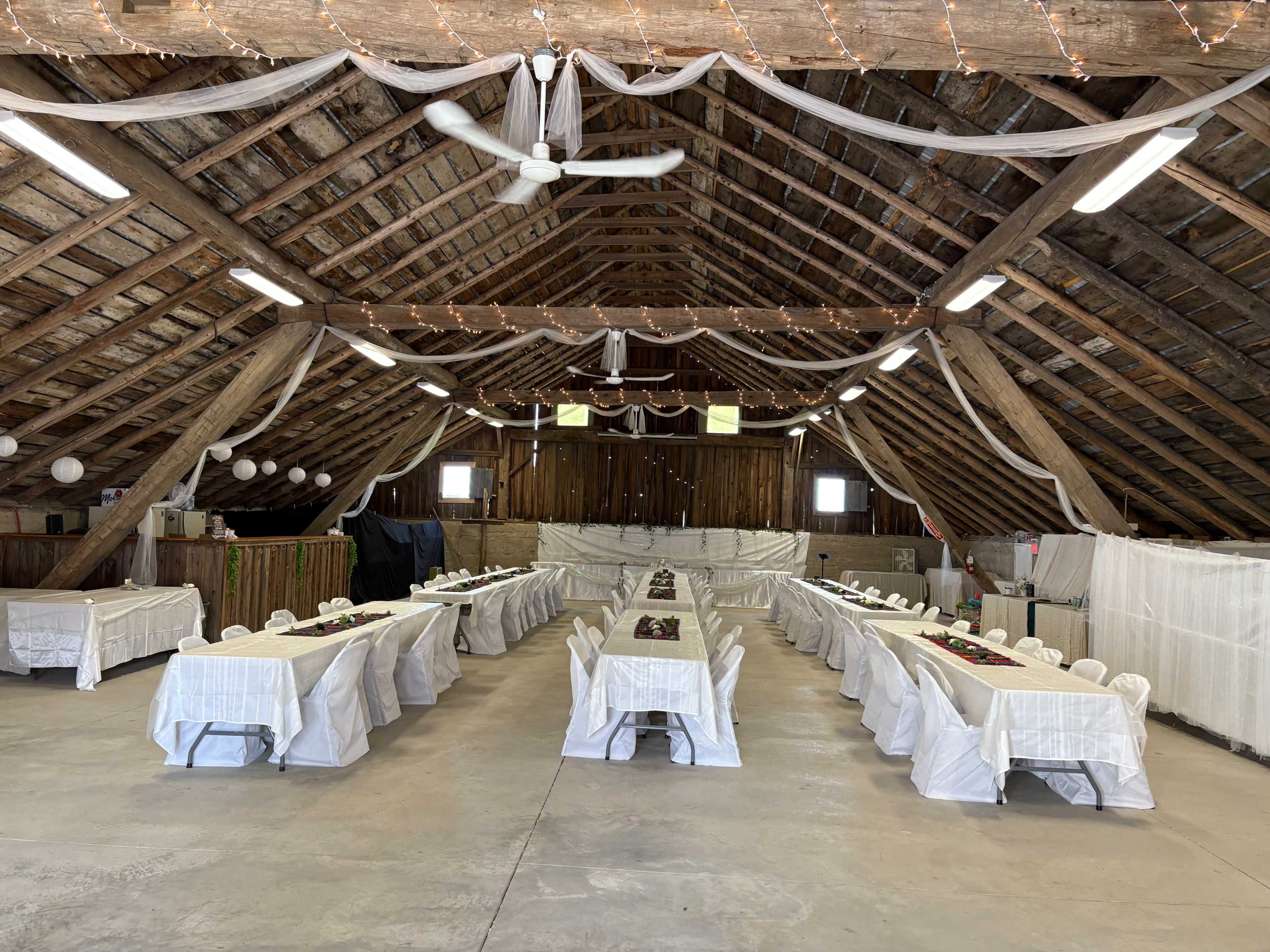 The image shows a large, rustic barn interior set up for an event, featuring tables covered with white tablecloths arranged in two long rows with chairs around them, and string lights and decorative draping overhead.
