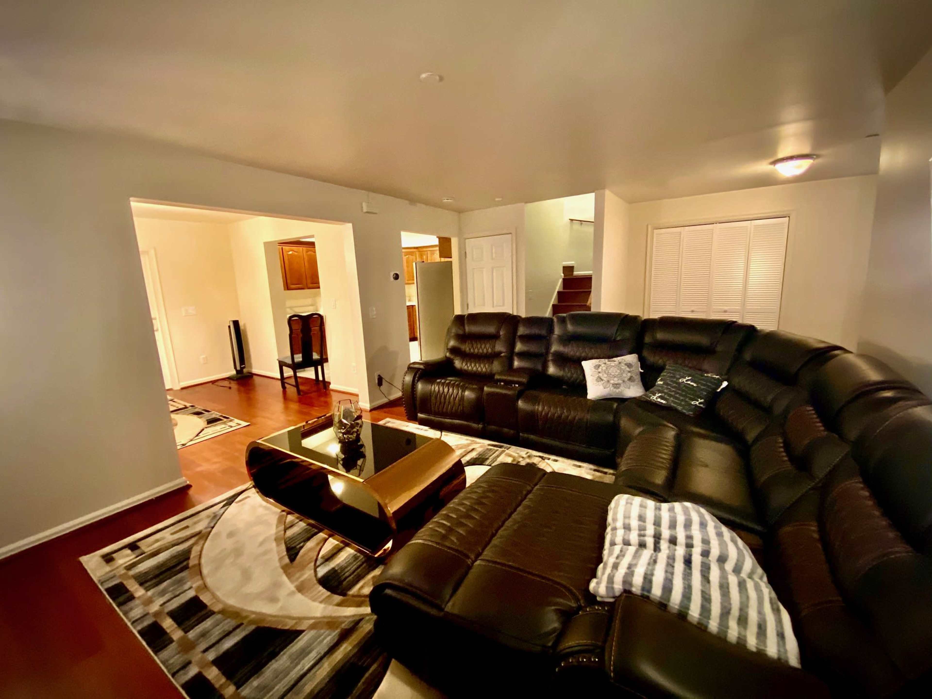 A spacious living room featuring a large black sectional sofa, a golden coffee table, and a patterned rug on the wooden floor.