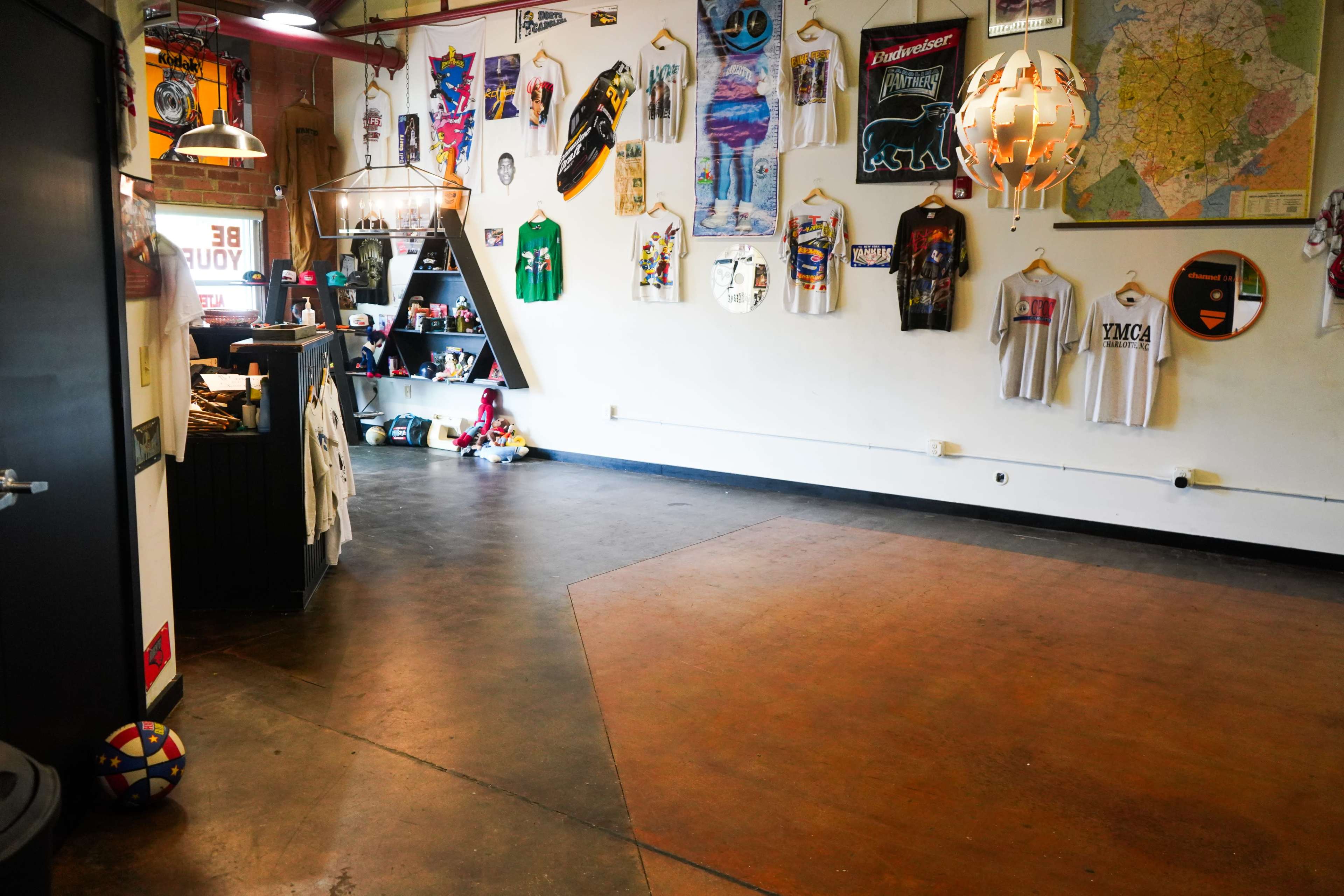 An interior space with a concrete floor, displaying various T-shirts on a wall and a small storefront area with shelves and a counter.
