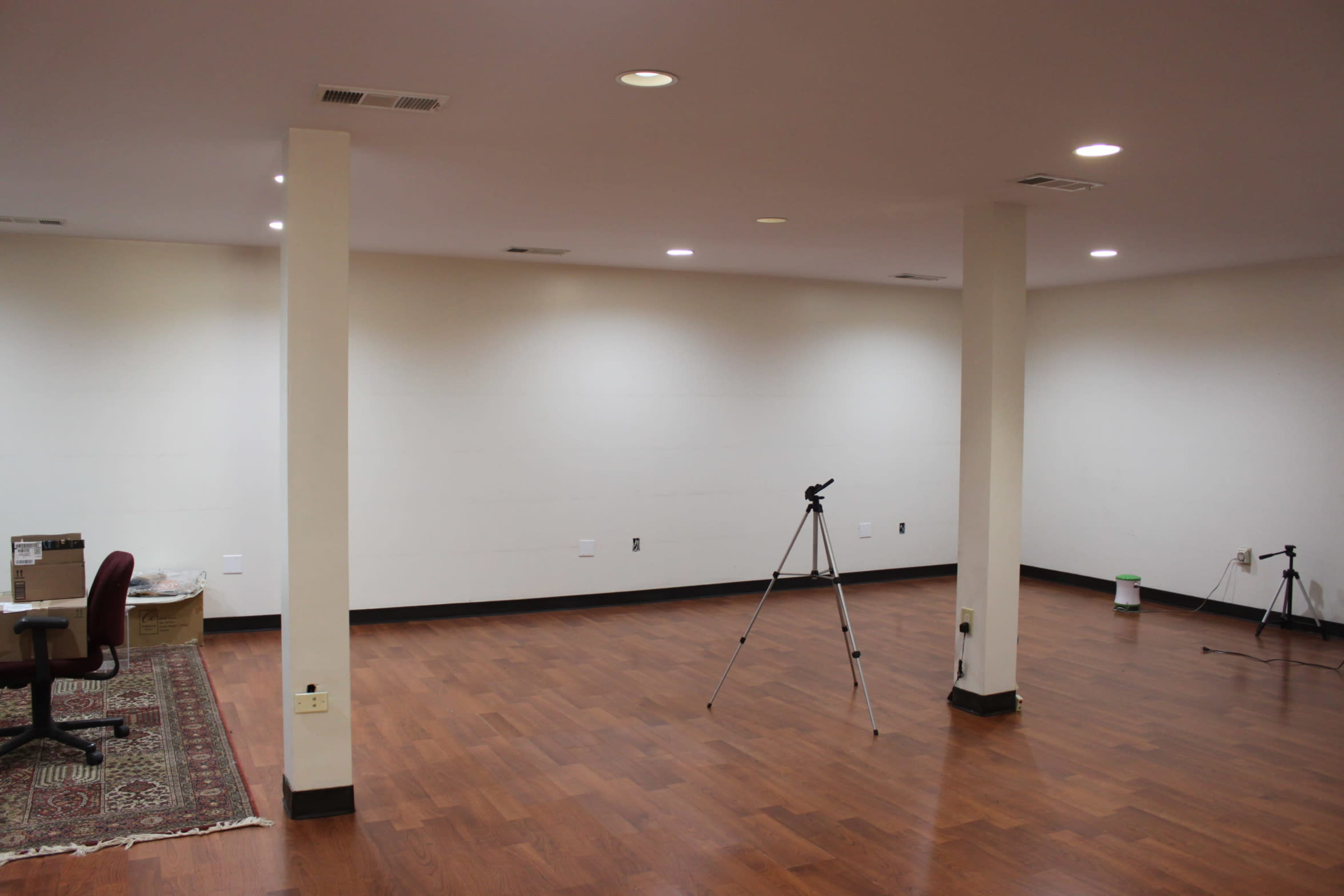 The image shows an empty room with wooden flooring, two columns, and a few tripods set up on one side.