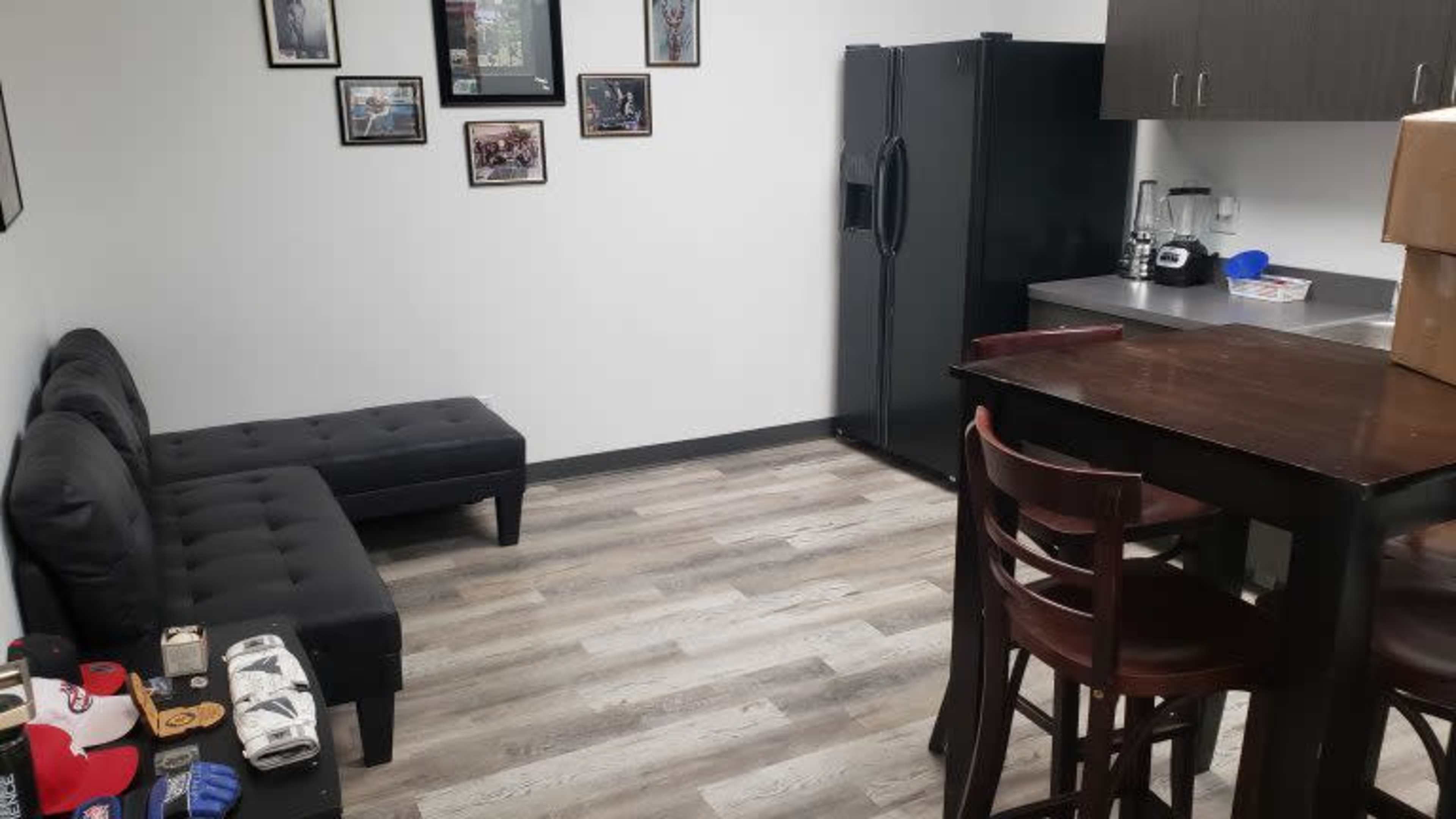 The image shows a small room with a black couch, a table with chairs, a refrigerator, and framed pictures on the walls.