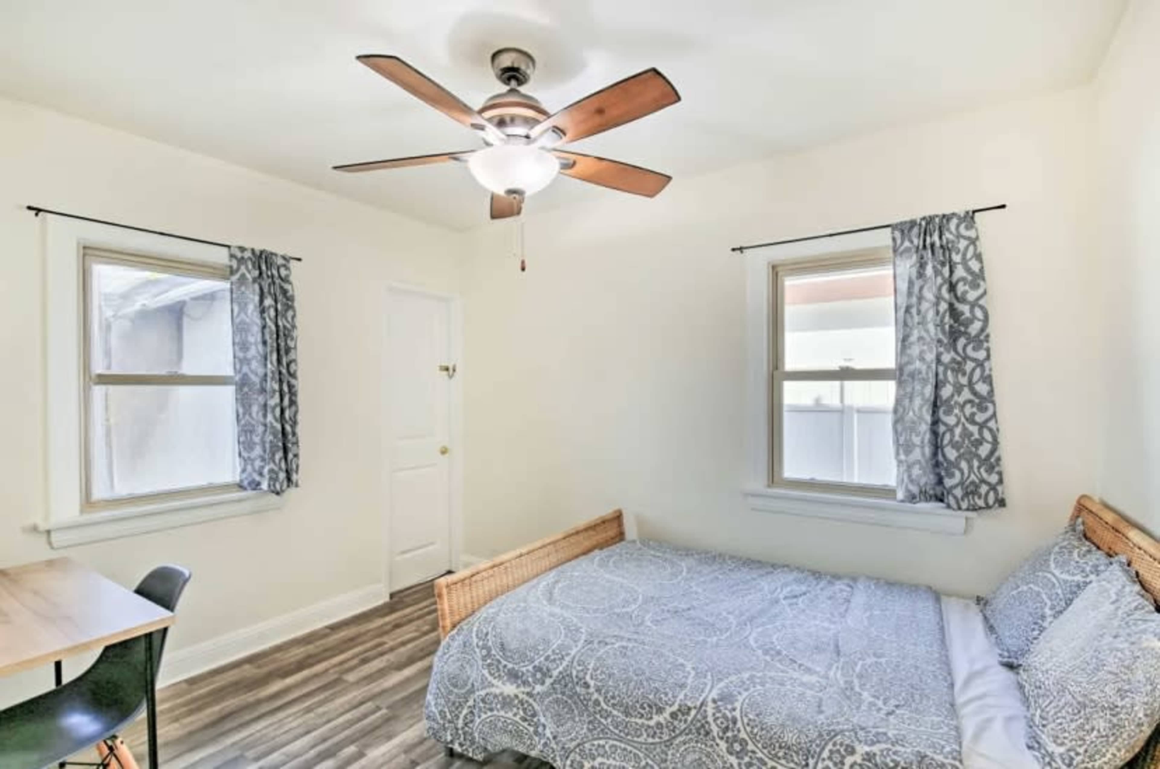 A small bedroom with a bed, a ceiling fan, a table with a chair, and two windows with curtains.