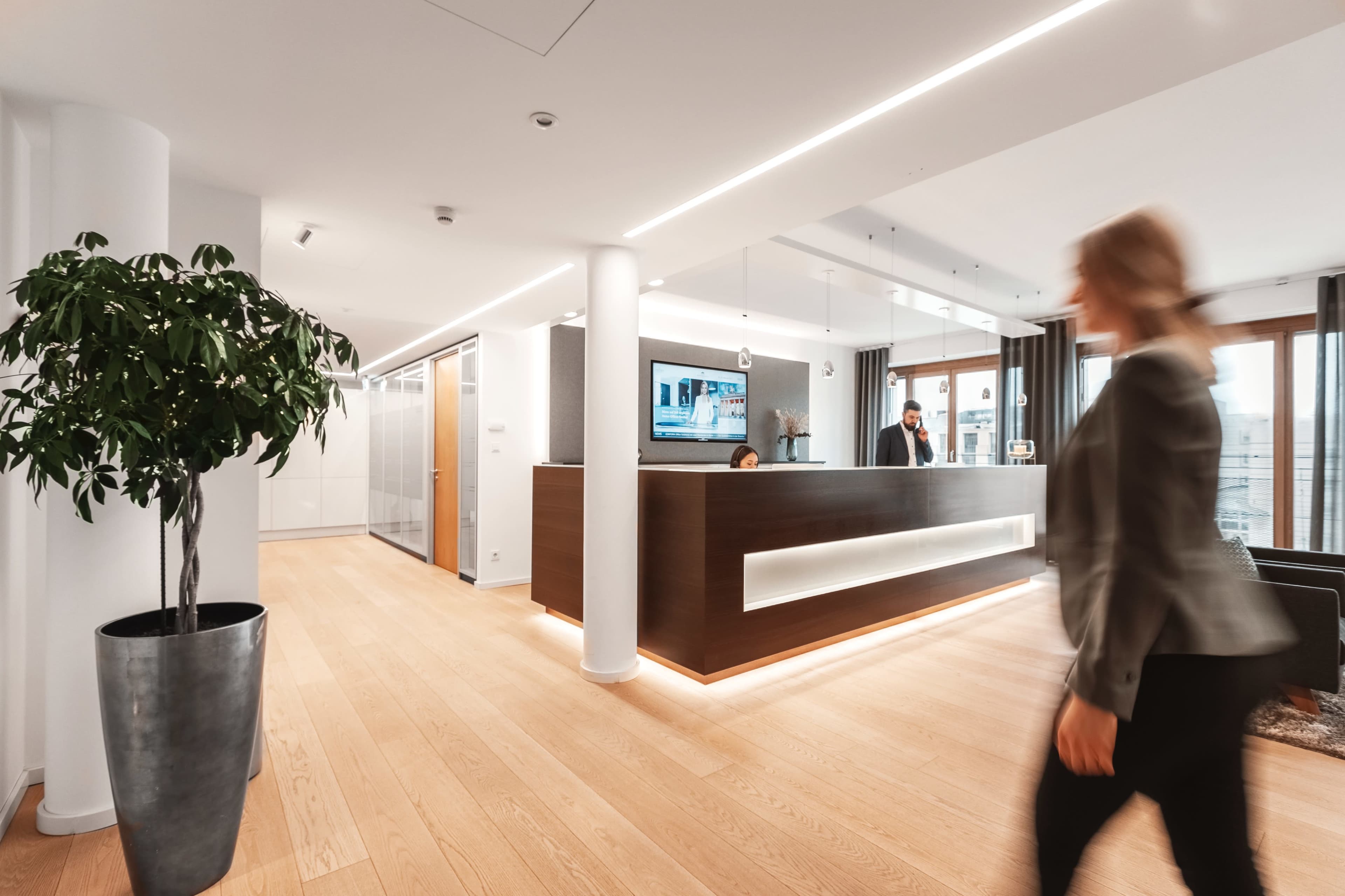A modern office reception area features a wood-paneled front desk, large windows with city views, and a person walking past a potted plant.