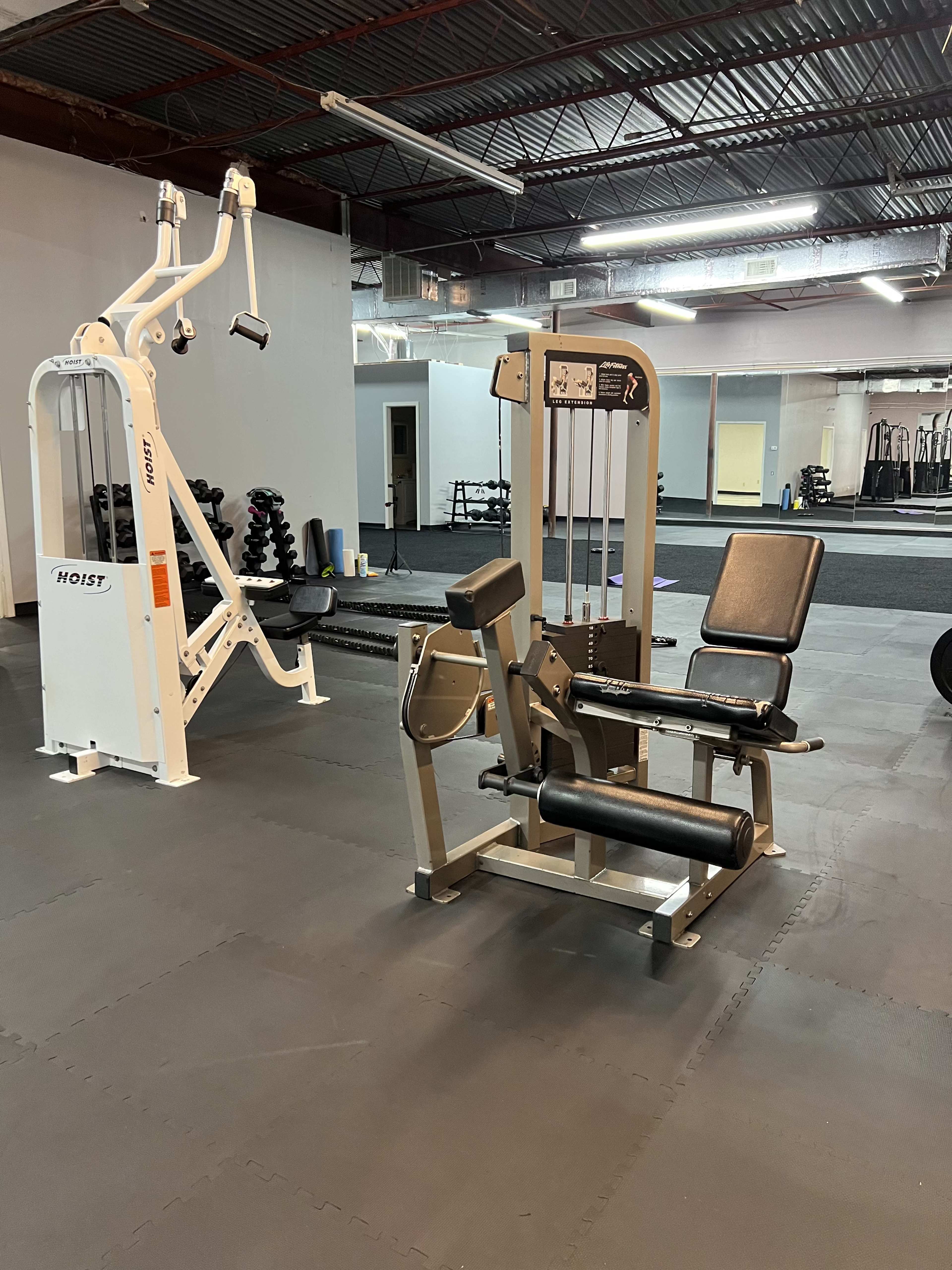 A fitness center interior featuring weight machines, including a Hoist machine on the left and a leg extension machine on the right, set on black rubber flooring.
