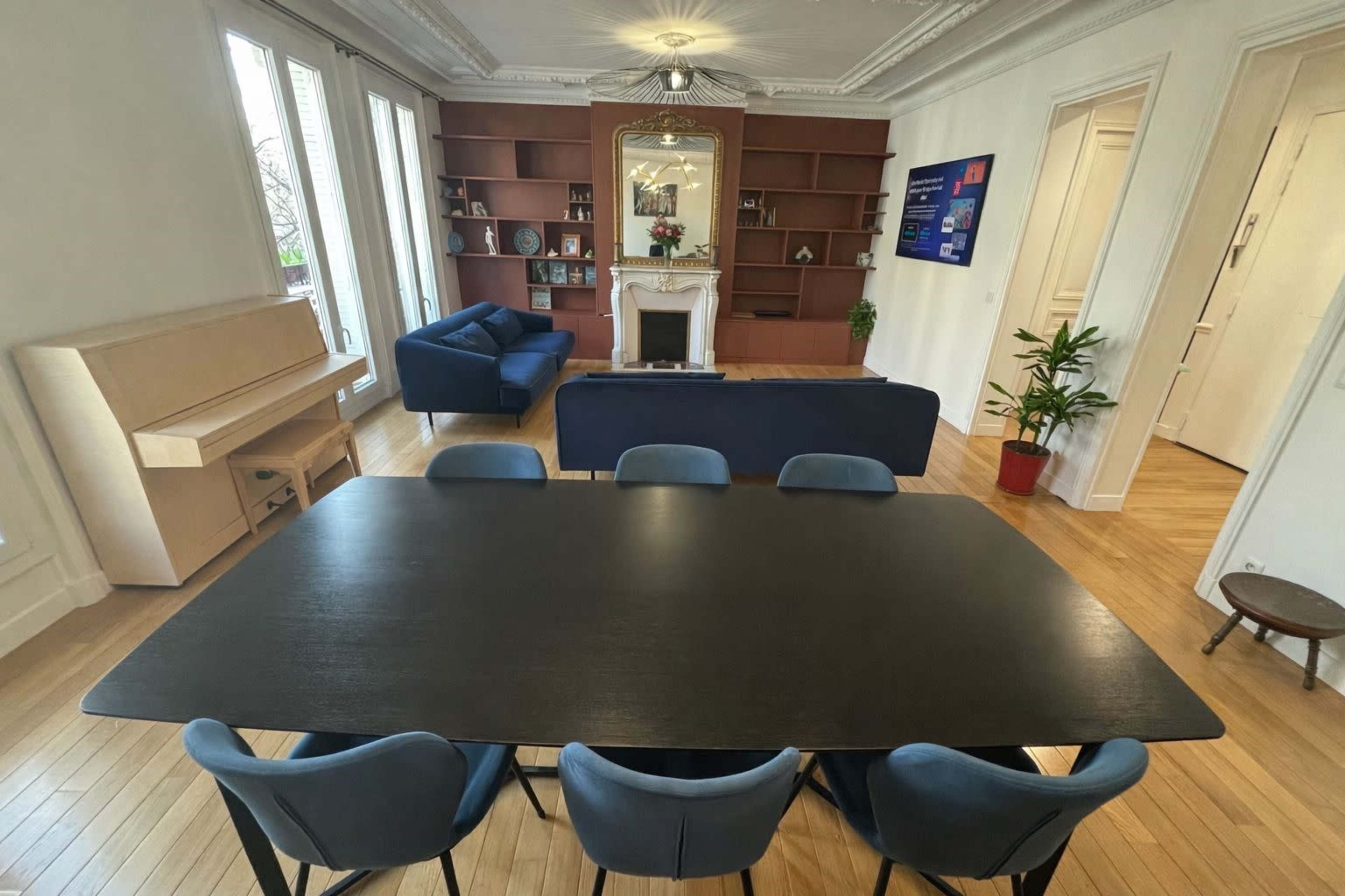 The image shows a spacious room featuring a large black dining table surrounded by blue chairs, with a sofa and a piano in the background.
