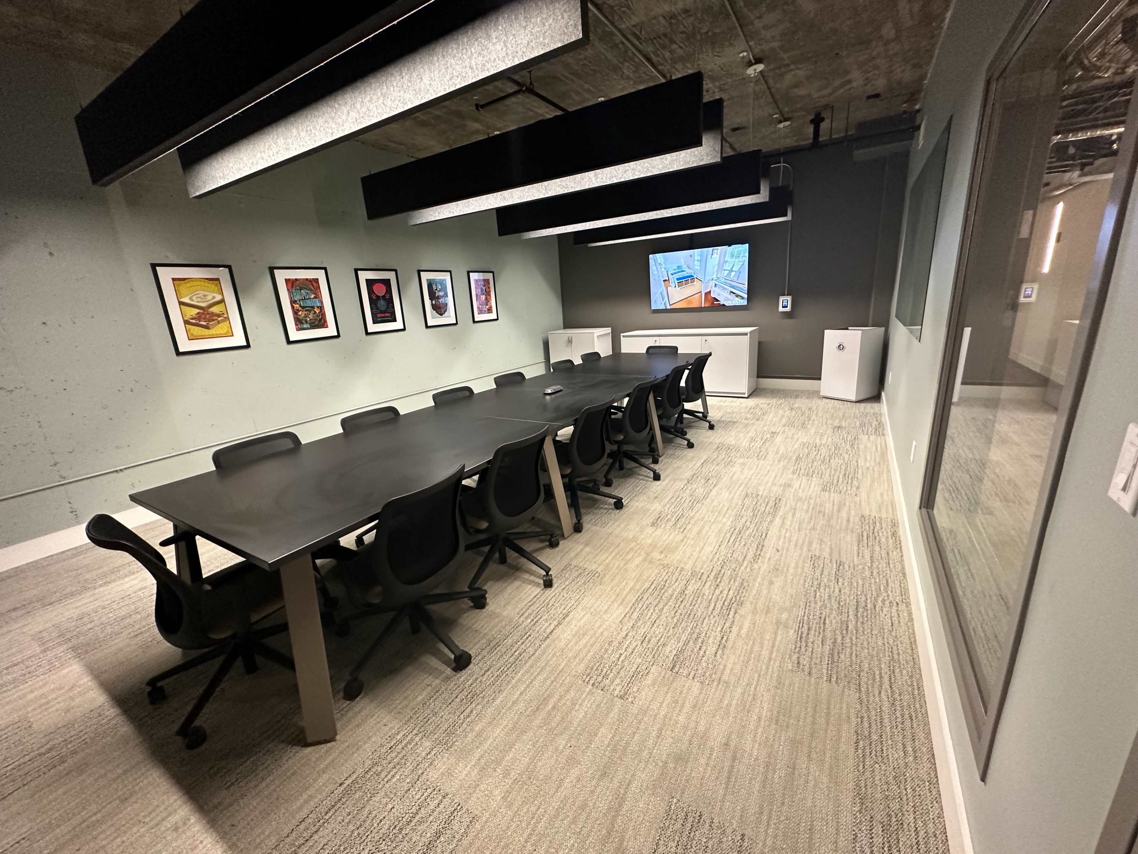 A conference room features a long table with black chairs, framed artwork on the walls, and a large screen at one end.