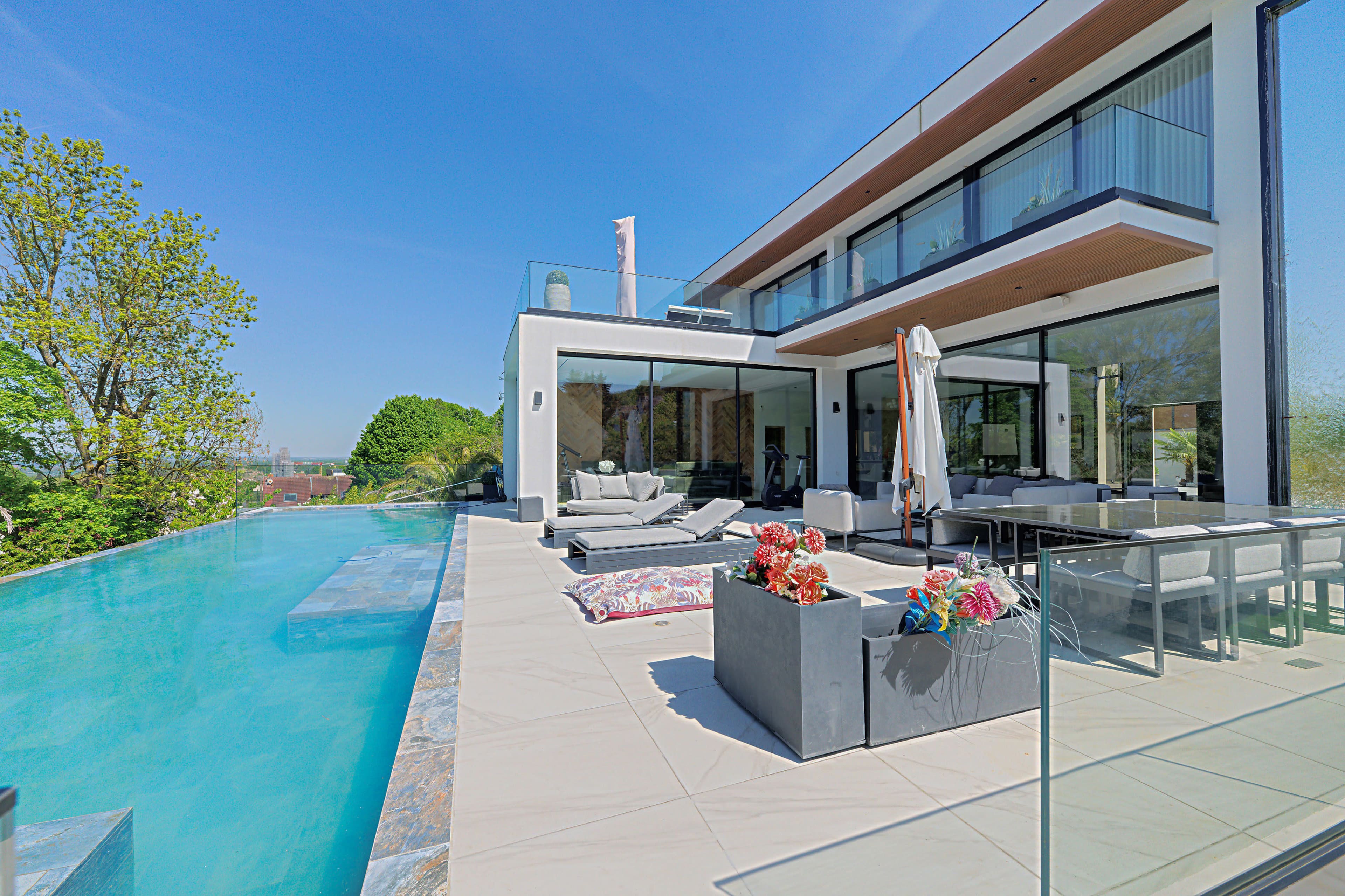 The image shows a modern house with a large infinity pool, outdoor seating area, and a well-maintained garden.