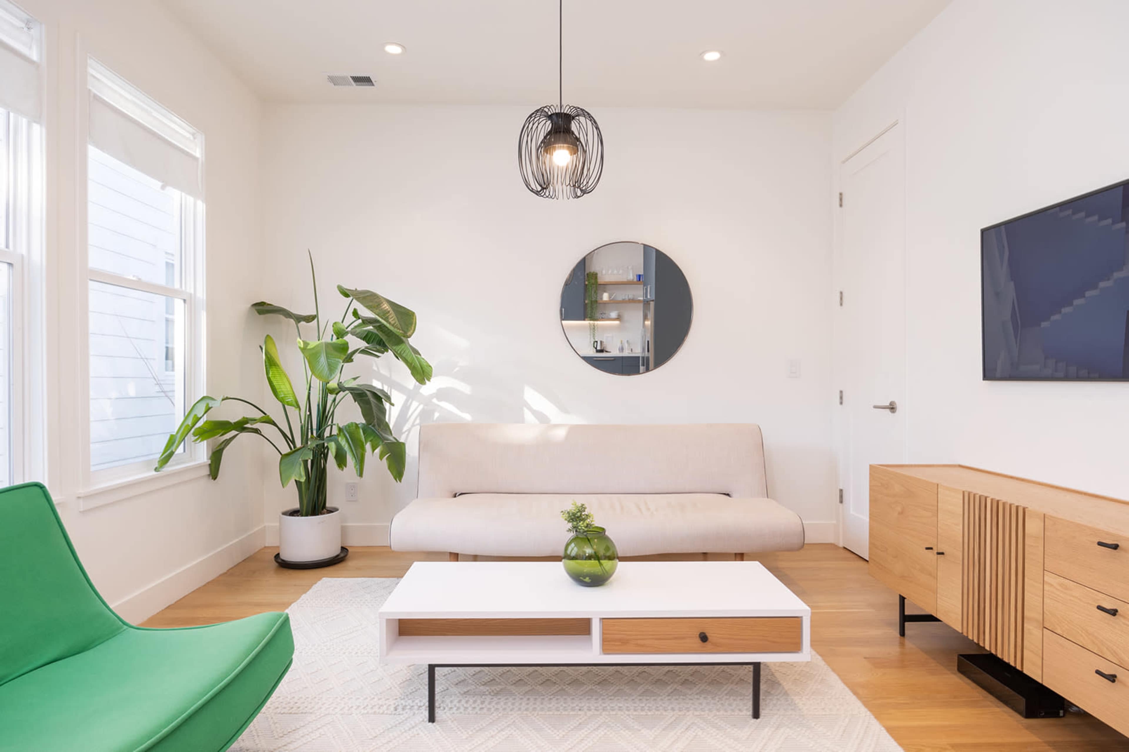 A modern living room features a light-colored sofa, a green lounge chair, a round mirror, and a wooden media console, complemented by a large indoor plant.