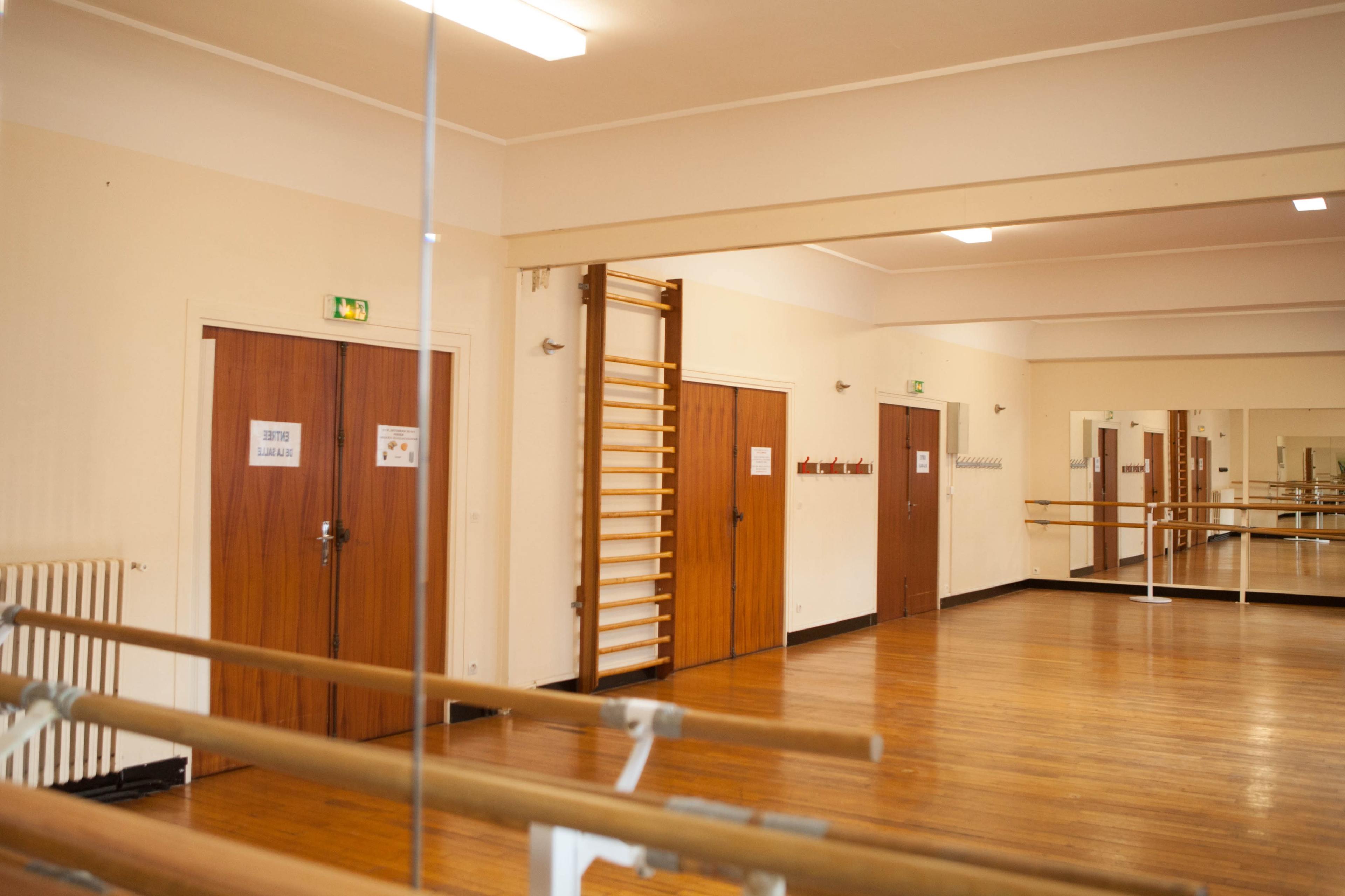 The image shows a spacious dance studio with wooden floors, mirrored walls, and several doors along one side.