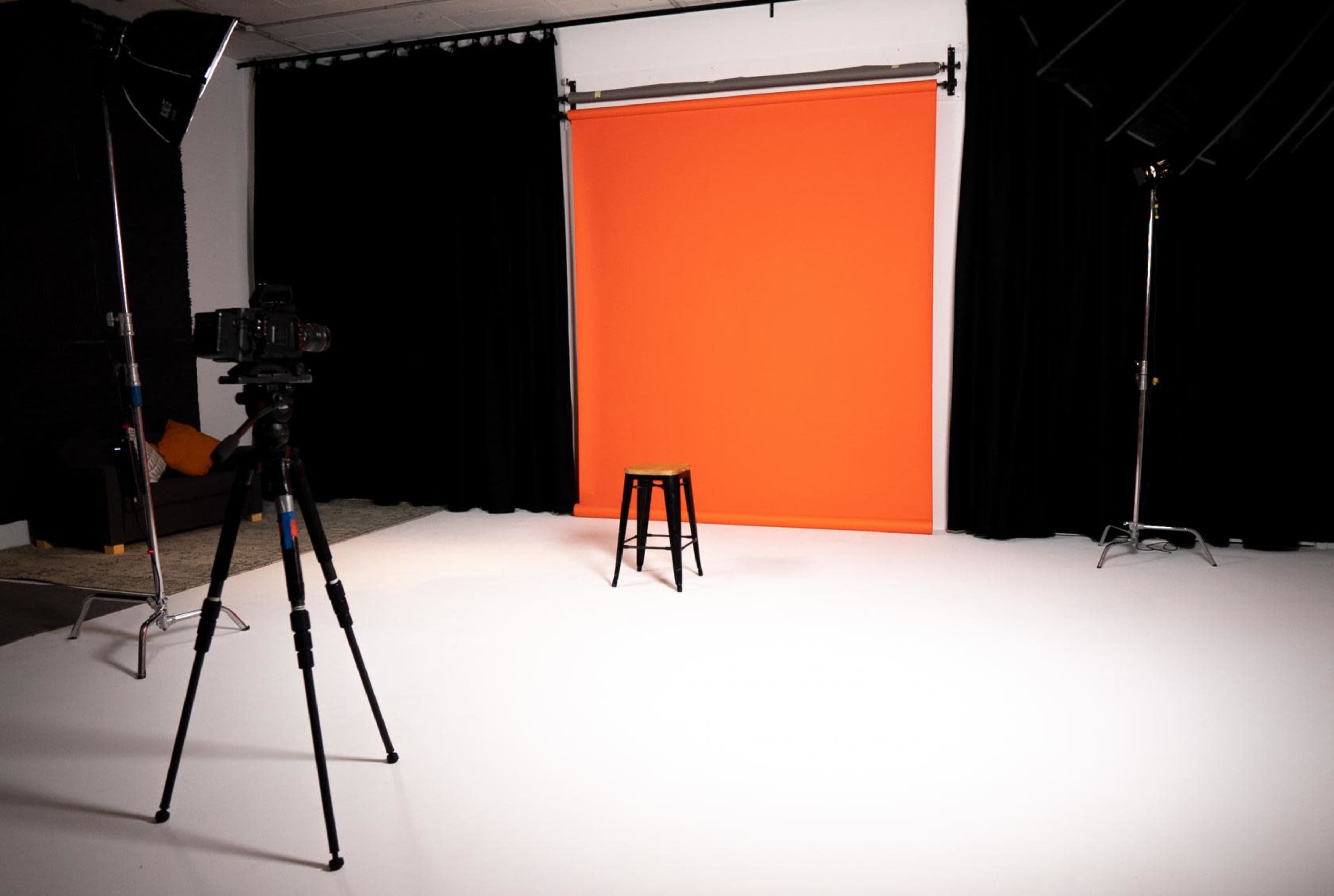 A camera and tripod face a black stool in front of an orange backdrop in a photography studio.
