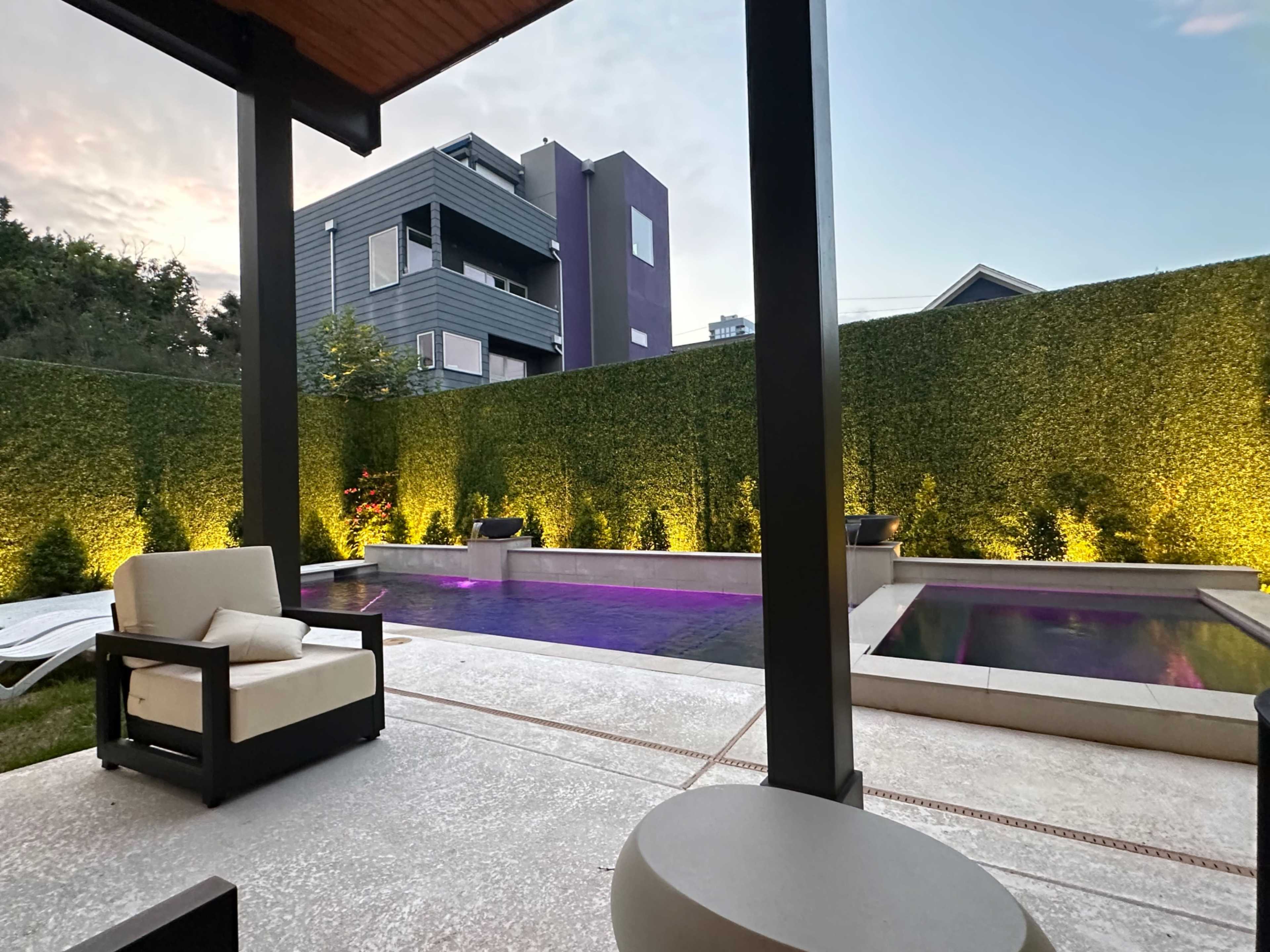 A modern outdoor space features a pool surrounded by tall green hedges, illuminated by soft lighting, with lounge chairs nearby.