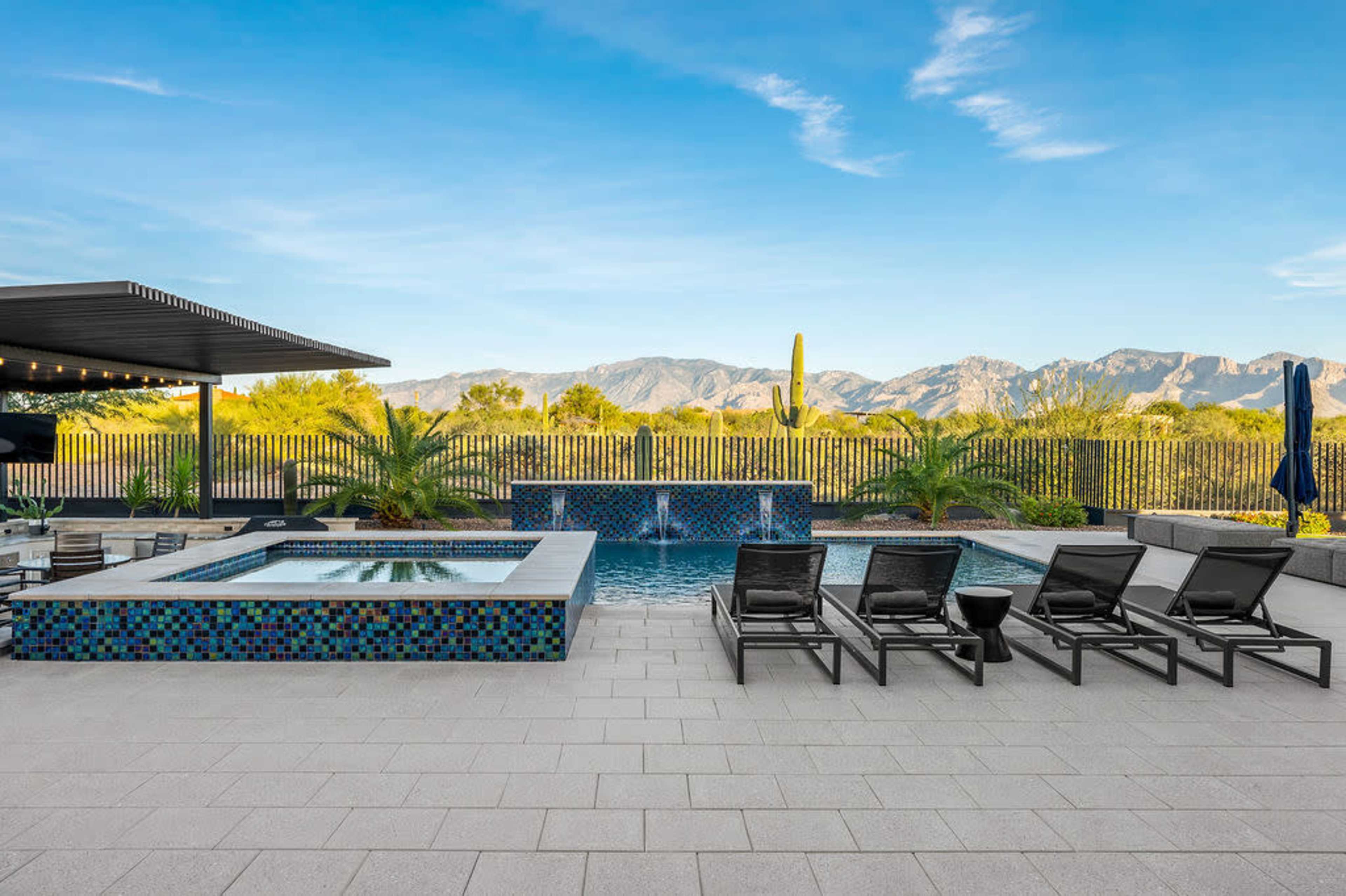 The image shows a modern outdoor pool area featuring a spa, lounge chairs, and a scenic view of mountains and greenery in the background.