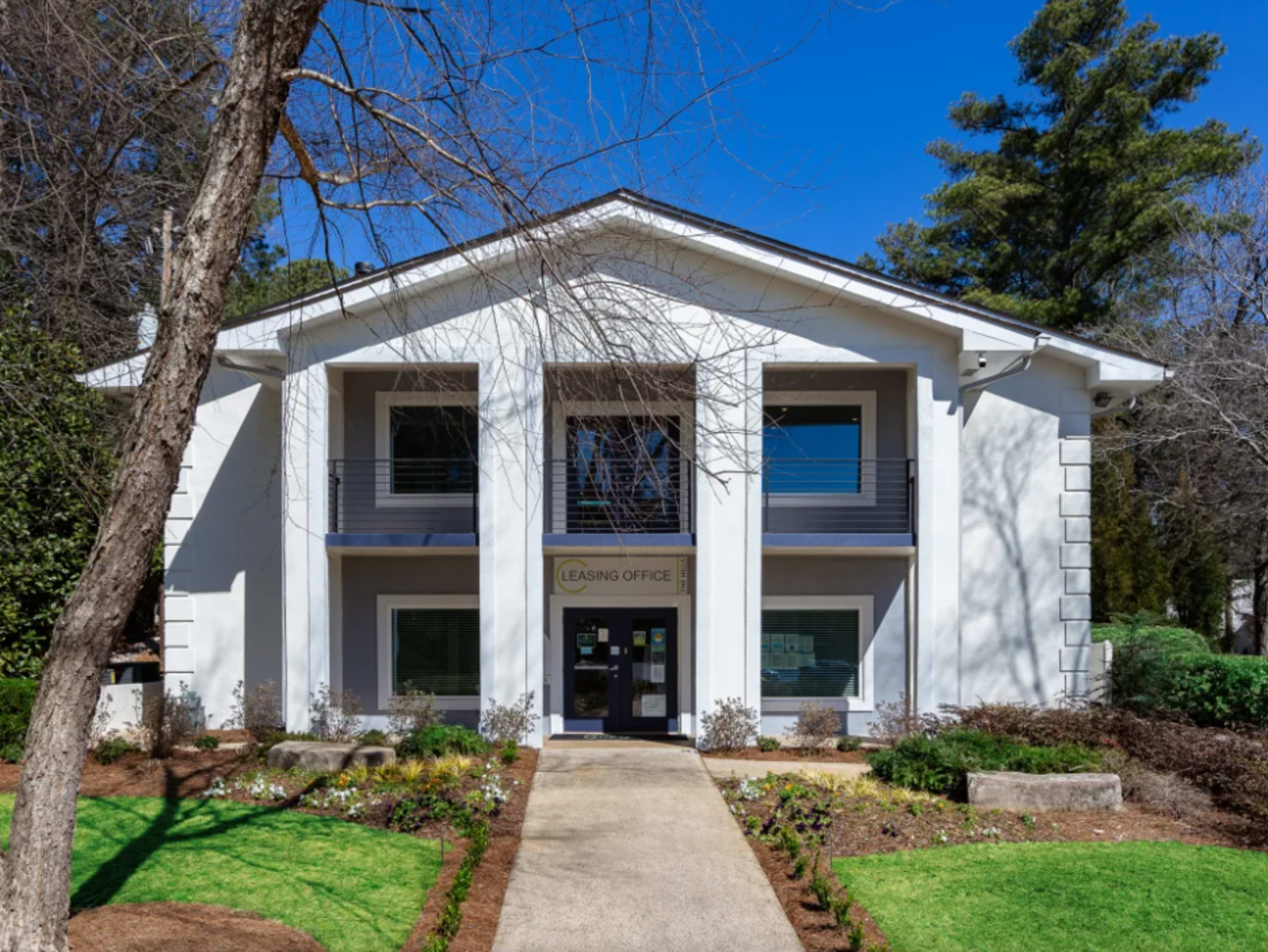 The image shows a two-story leasing office building with a landscaped pathway and greenery surrounding it.