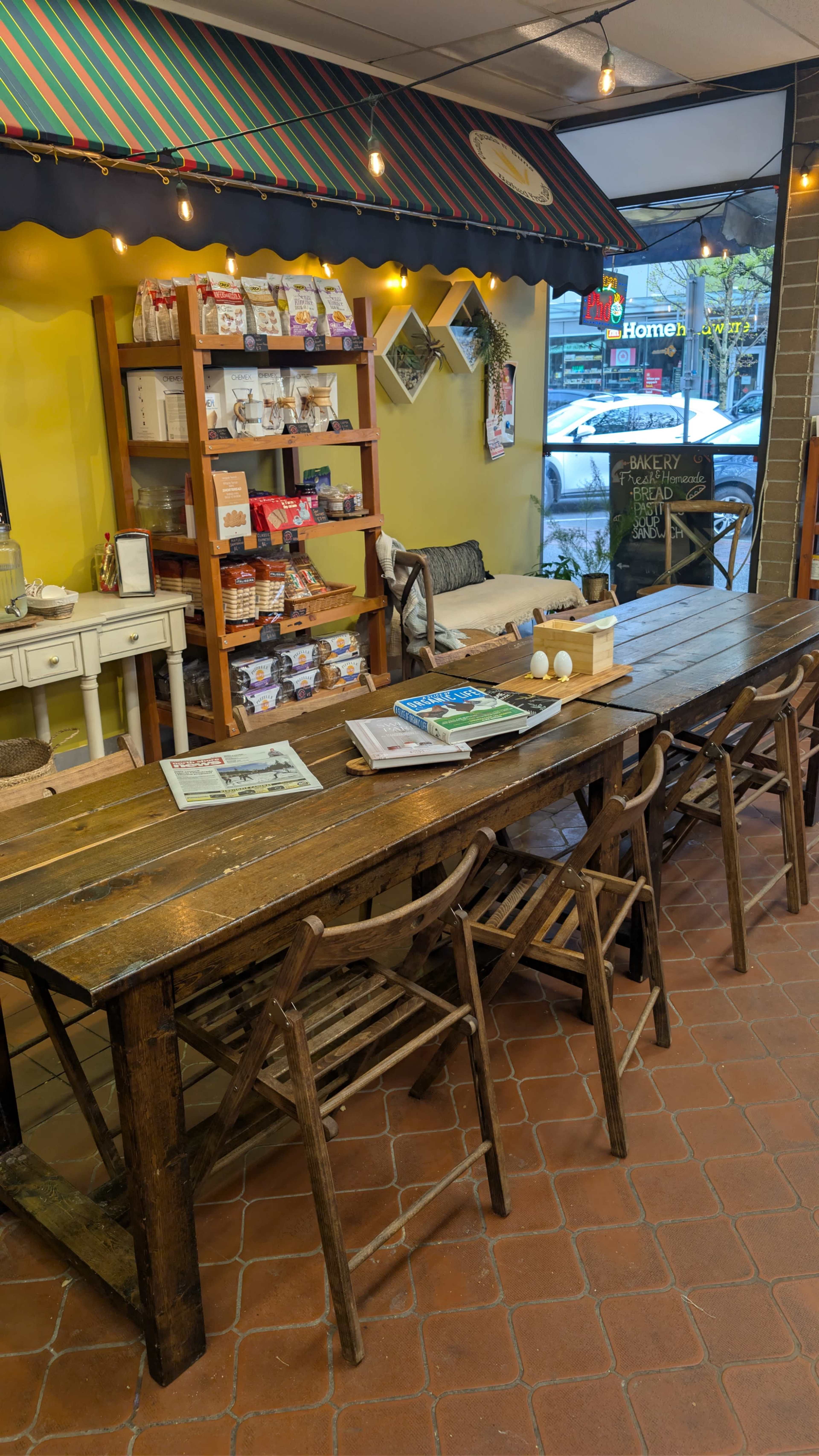 A large wooden table with chairs surrounds it, set in a brightly lit café featuring shelves stocked with various products along the wall.