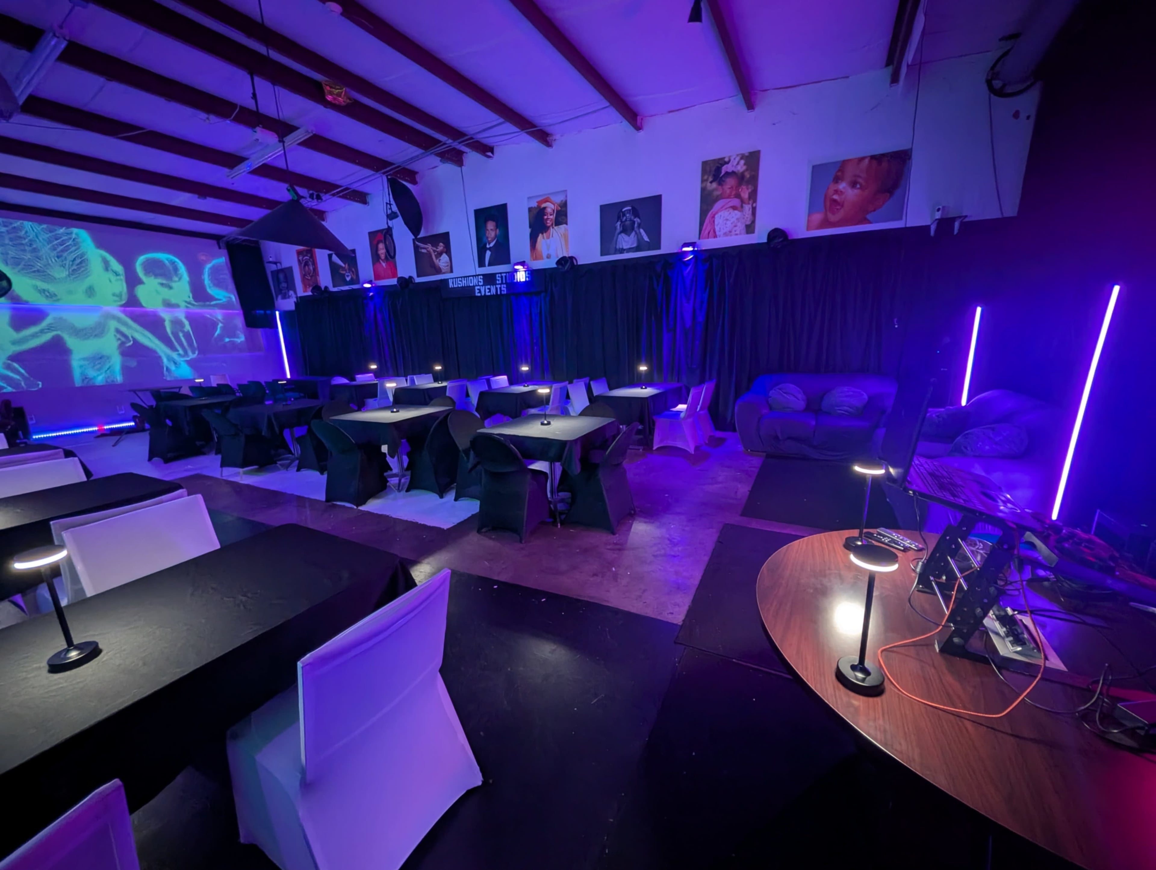 The image shows a dimly lit event space with black tables and chairs arranged for a gathering, colorful lighting, and various artworks displayed on the walls.