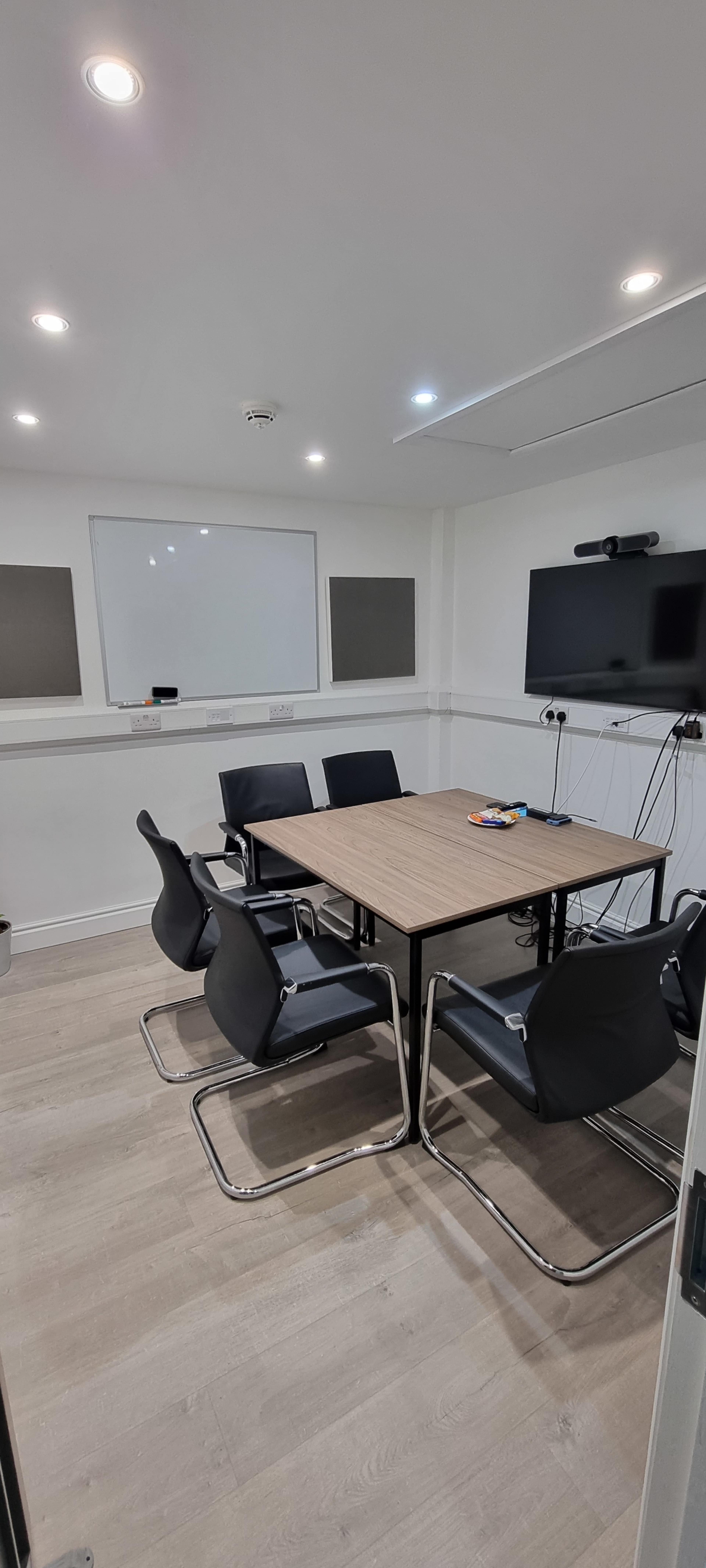 A small conference room features a rectangular table surrounded by six black chairs, a whiteboard, and a television mounted on the wall.
