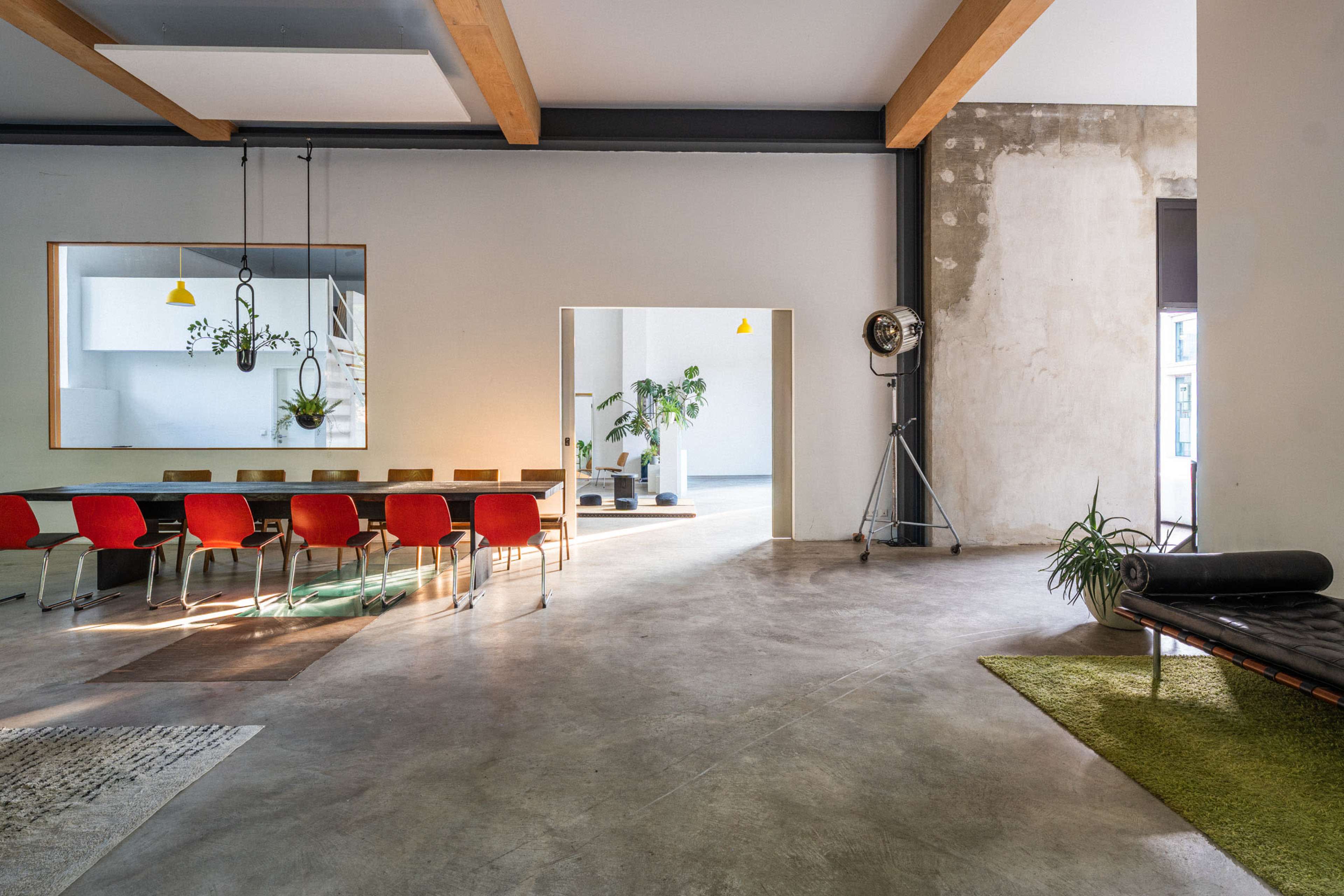 The image shows a spacious, modern interior with a long dining table and red chairs, featuring wooden beams and a mix of concrete and plaster walls.