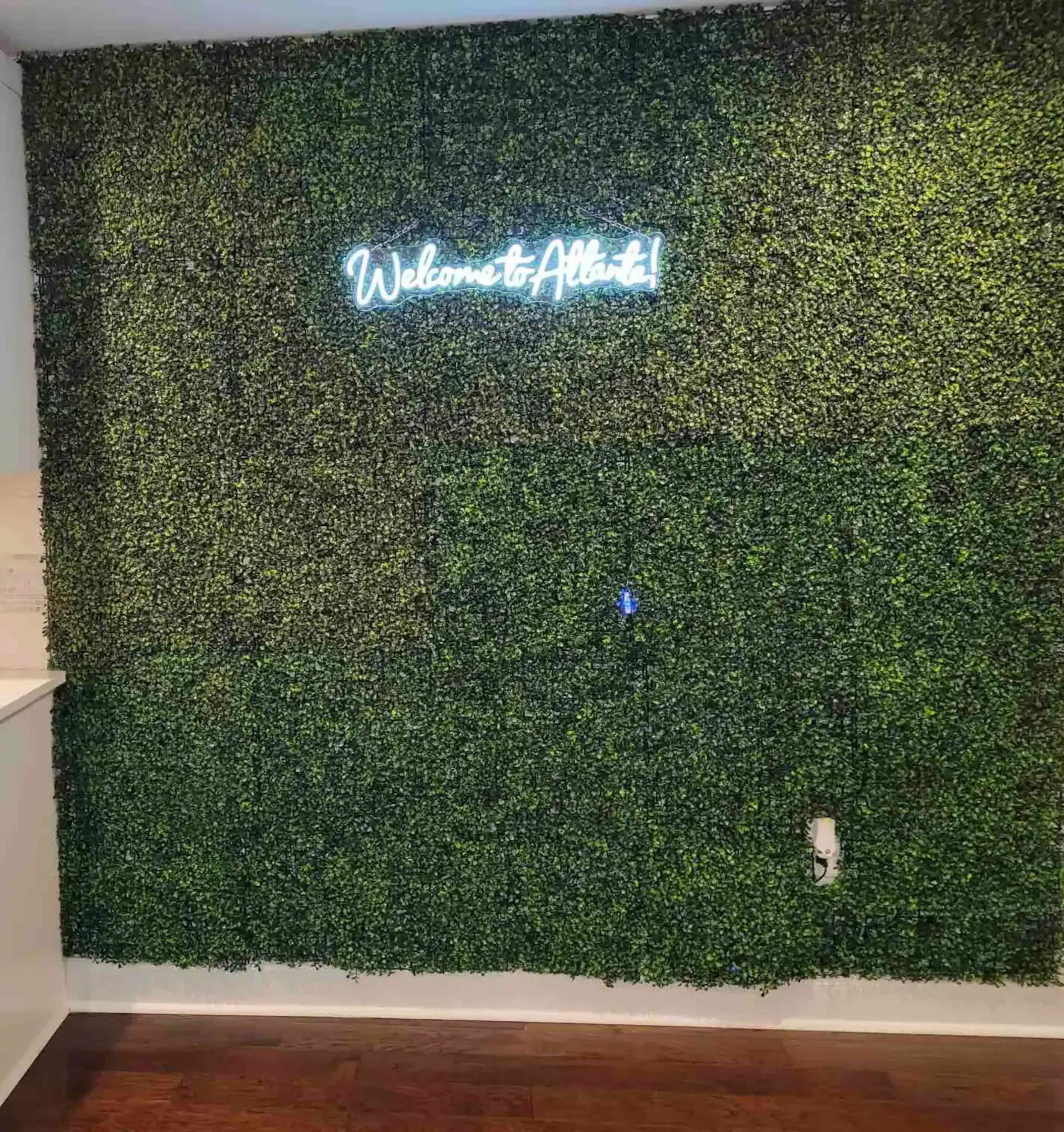 A green wall covered in artificial foliage displays a neon sign that reads "Welcome to Atlanta!"