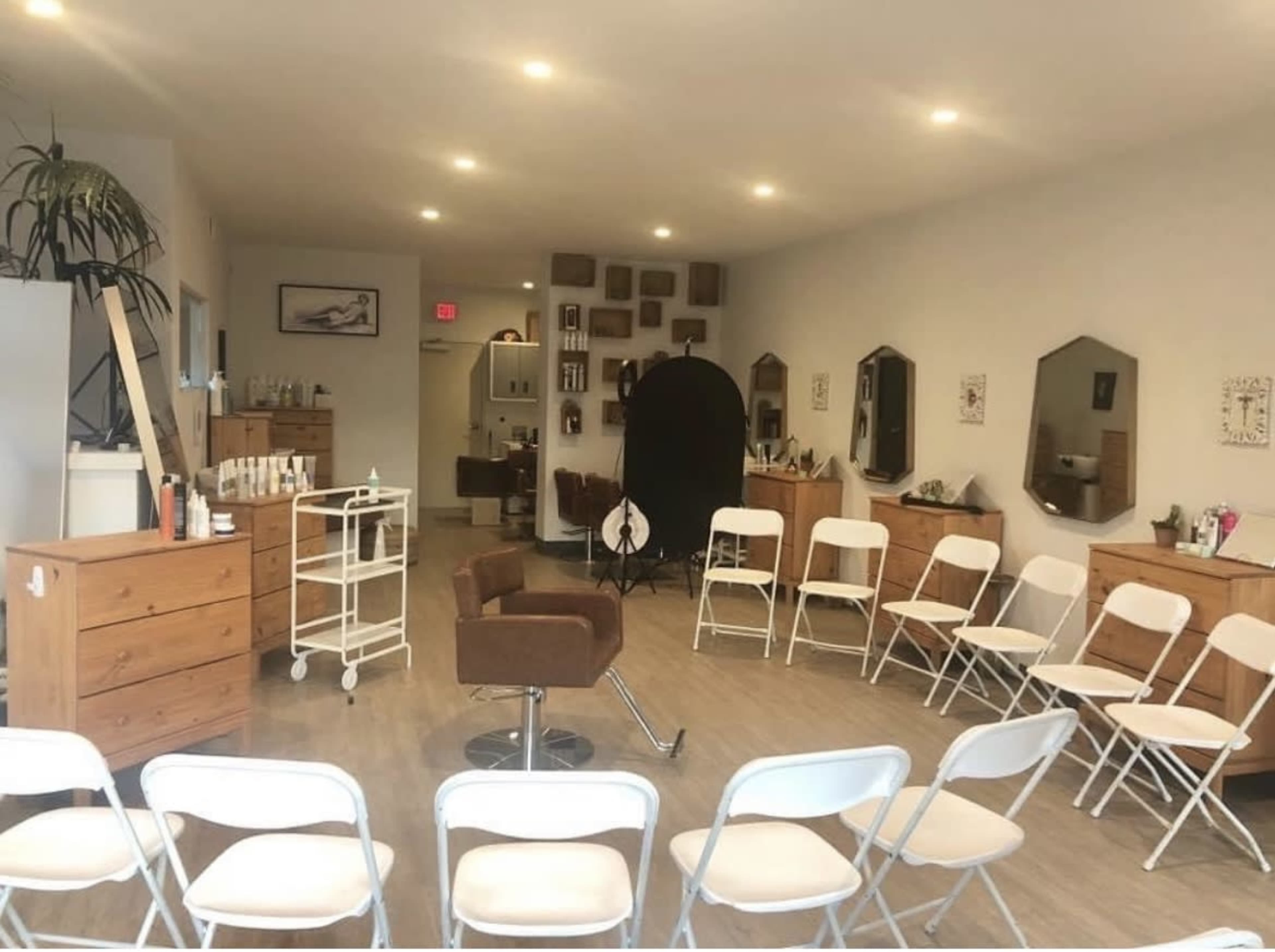 A hair salon interior with wooden dressers, a styling chair in the center, and white folding chairs arranged in a semi-circle.