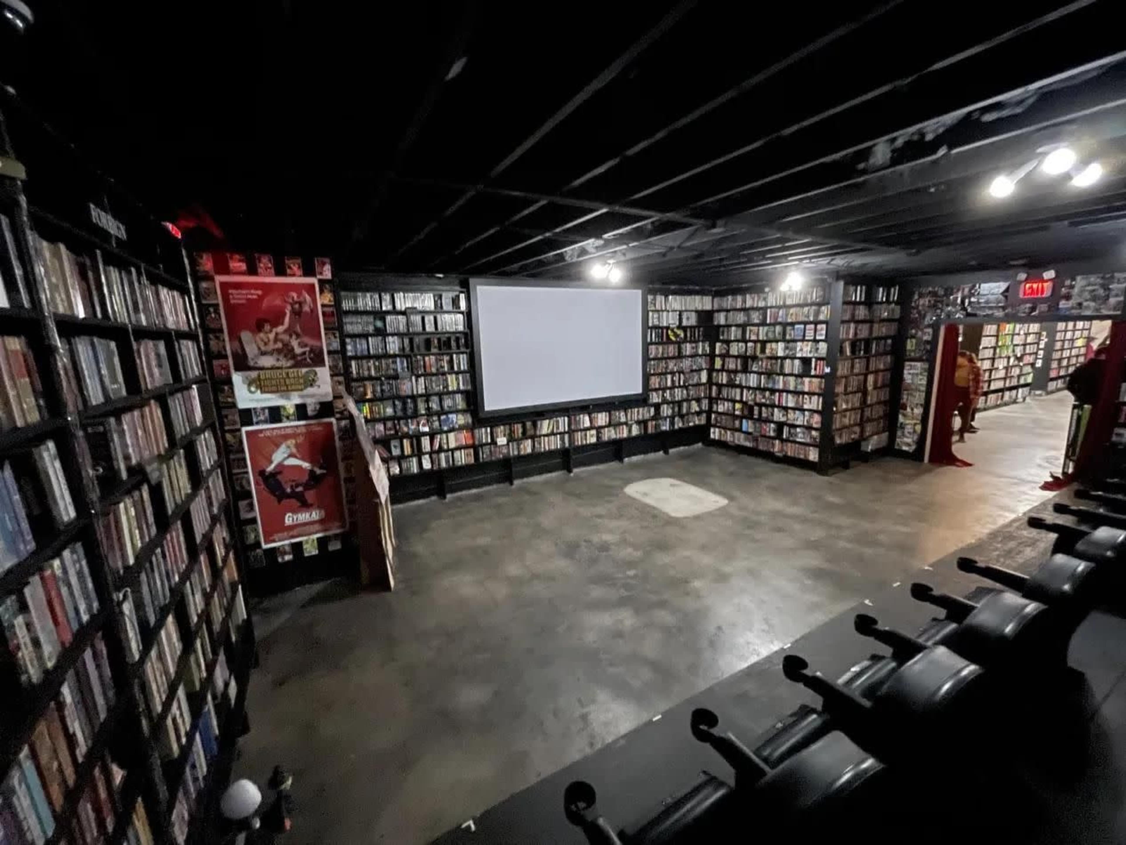 The image shows a dimly lit room filled with shelves of DVDs lining the walls, featuring a large projection screen at one end.