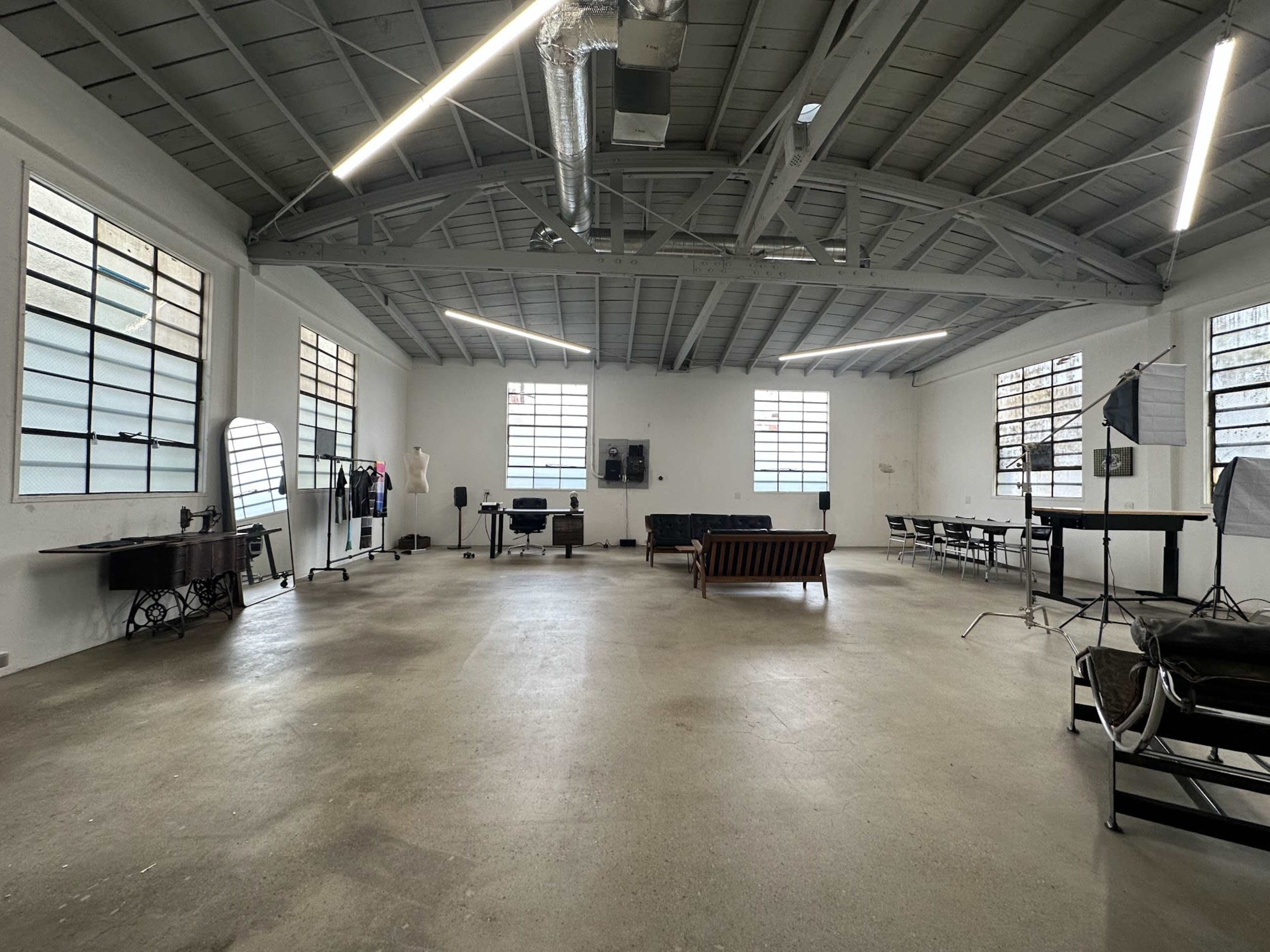 A spacious, well-lit studio with large windows, featuring various furniture pieces and photography equipment arranged around the room.