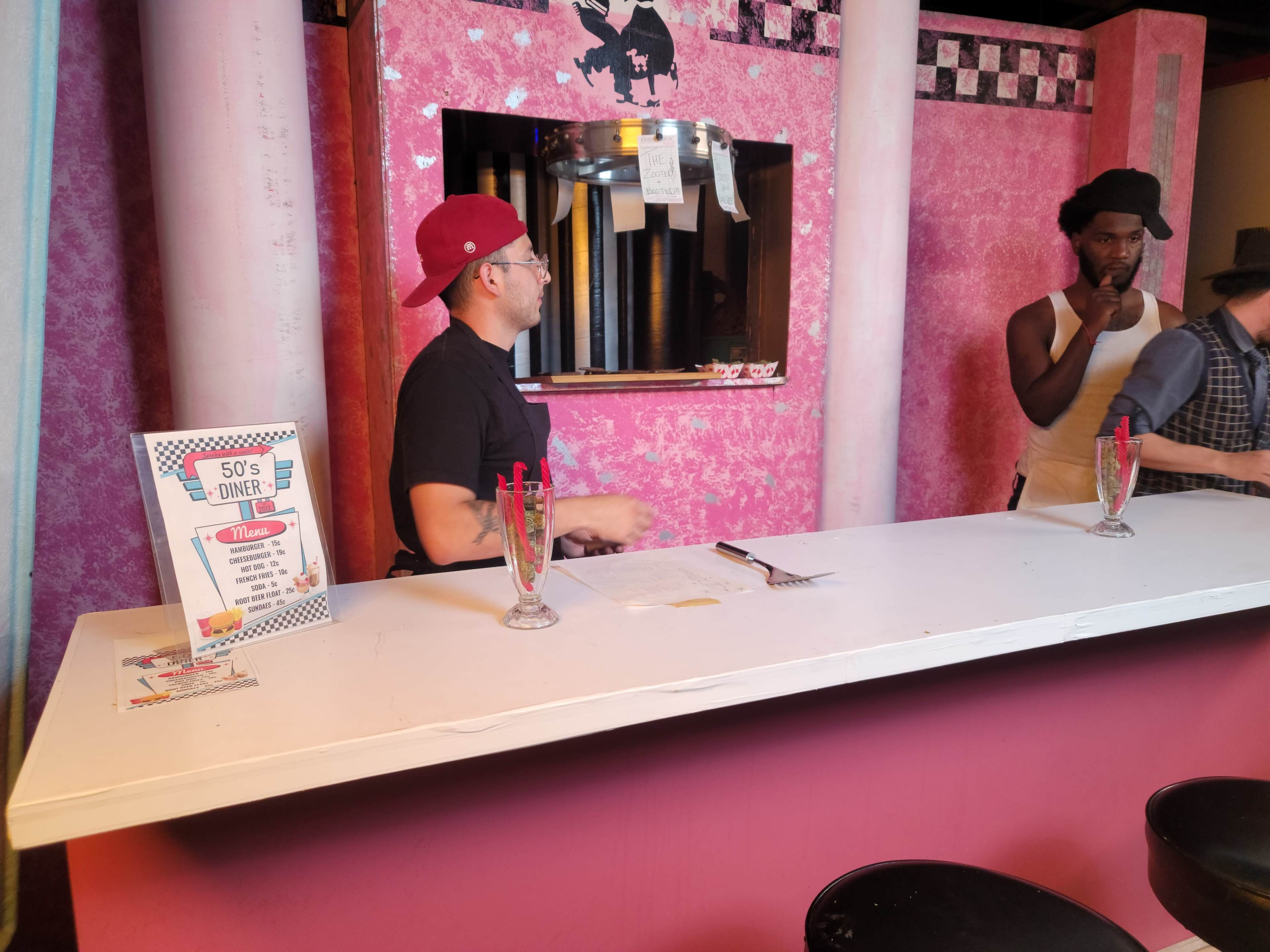 A countertop at a retro-style diner is adorned with milkshake glasses, while two men, one wearing a cap and the other in period attire, stand behind the bar.