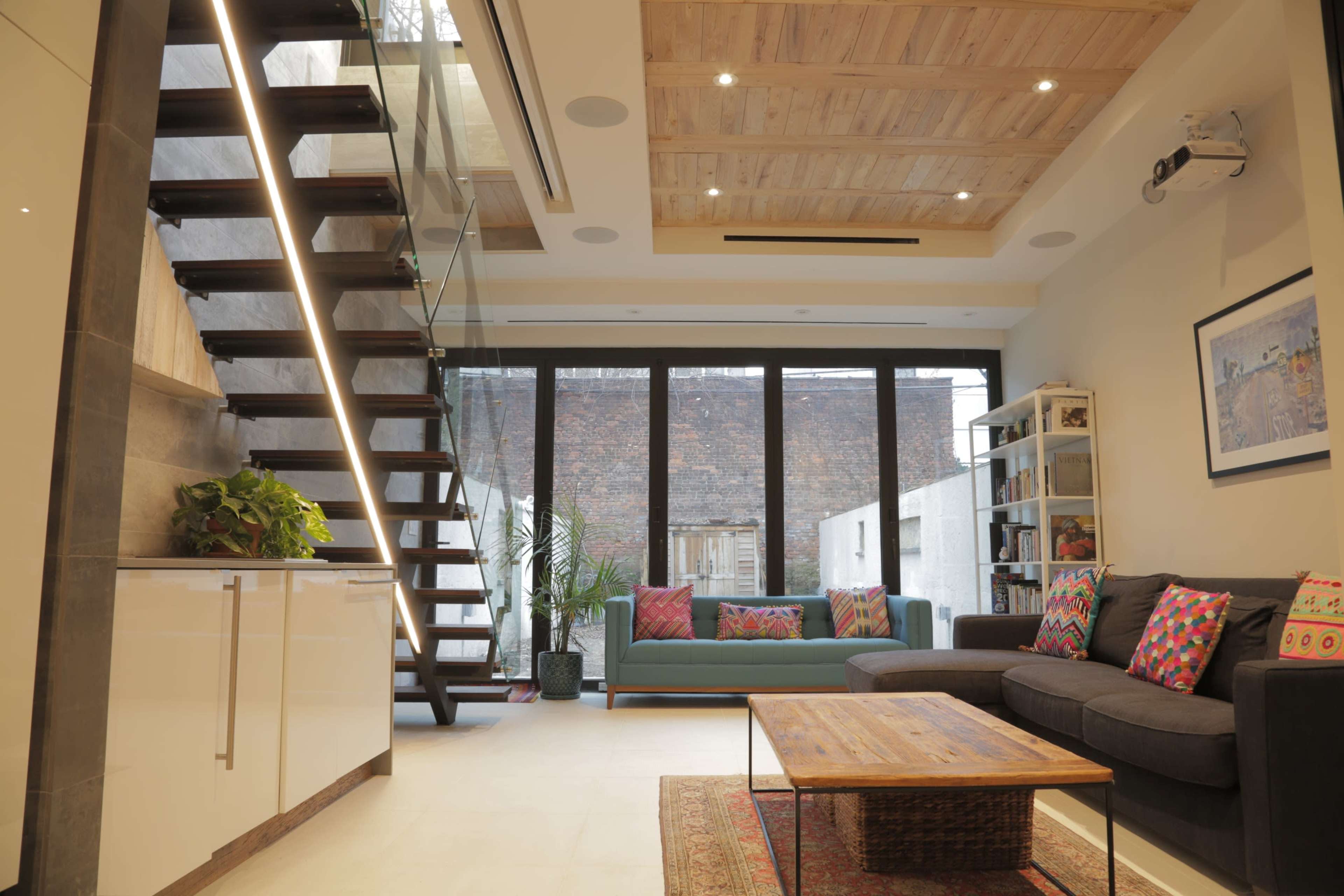 The image shows a modern living room with a glass staircase, a couch adorned with colorful cushions, and a wooden coffee table, set against a backdrop of large windows.