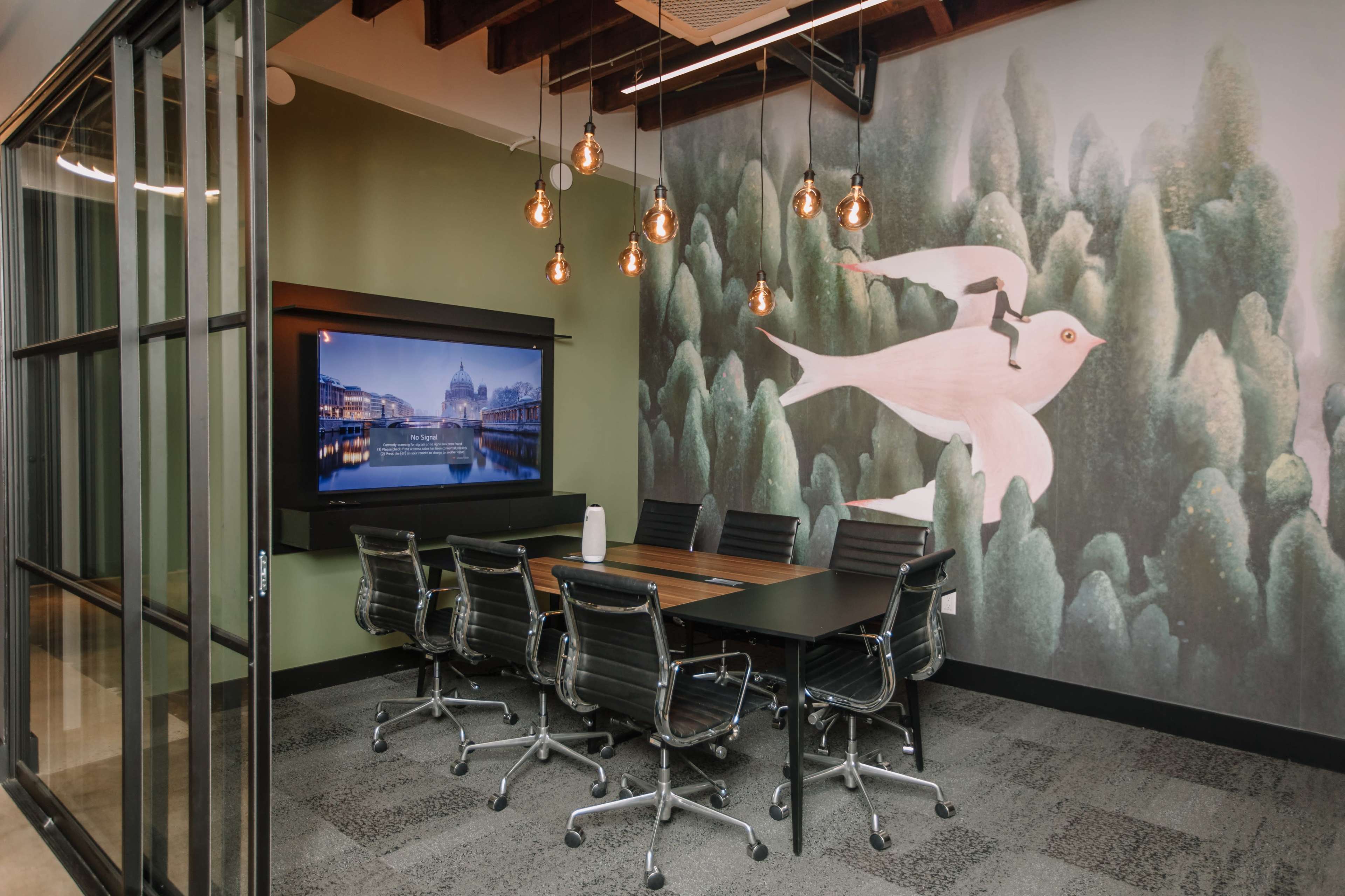 The image shows a modern conference room with a large screen on one wall and a mural of birds and trees on the opposite wall, surrounded by dark office chairs and a wooden table.