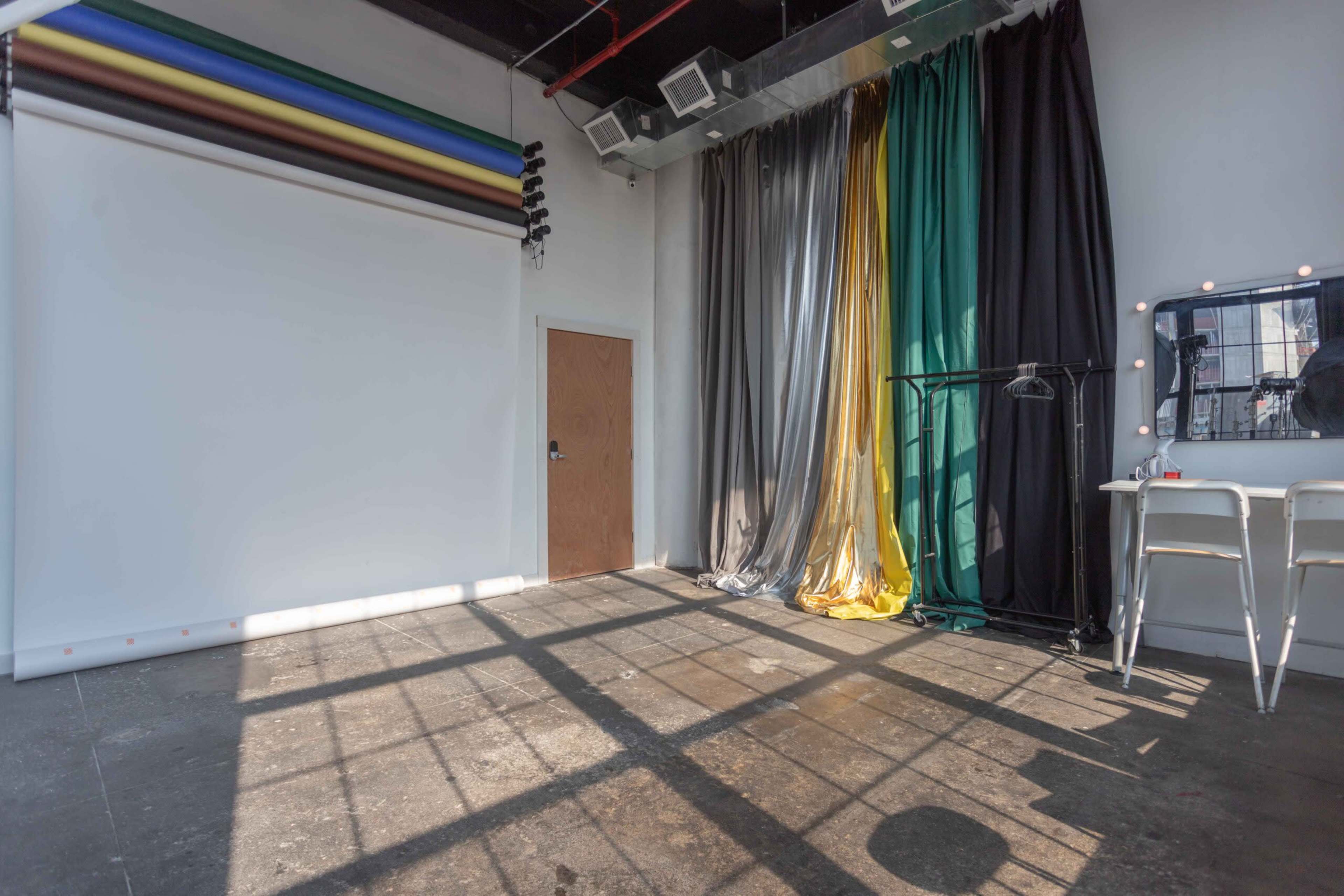 The image shows a photography studio with a white wall, colorful backdrop rolls, metallic curtains, and a mirror with light bulbs.