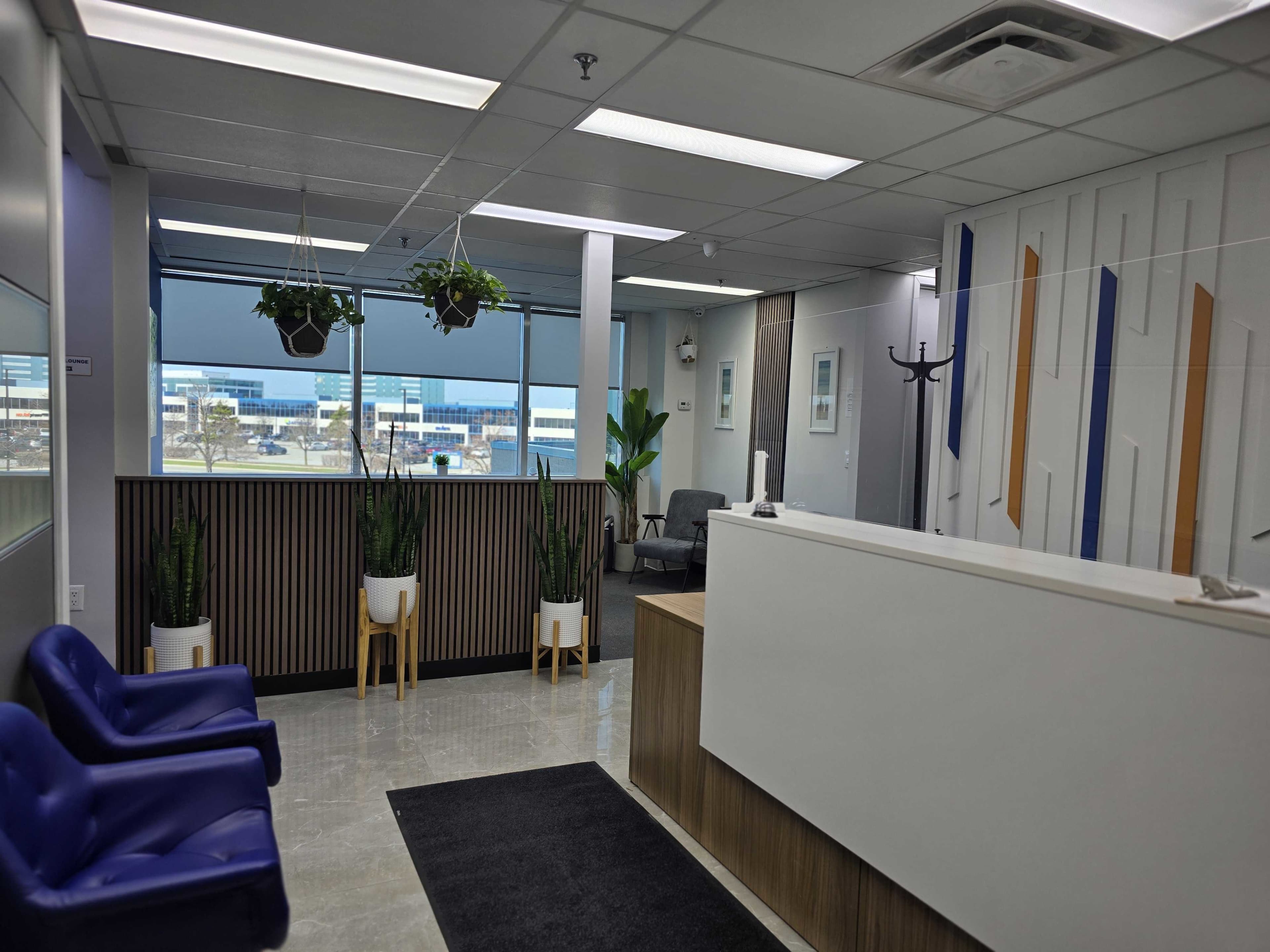 The image shows a modern reception area with a front desk, seating, and indoor plants, featuring large windows that overlook an exterior landscape.