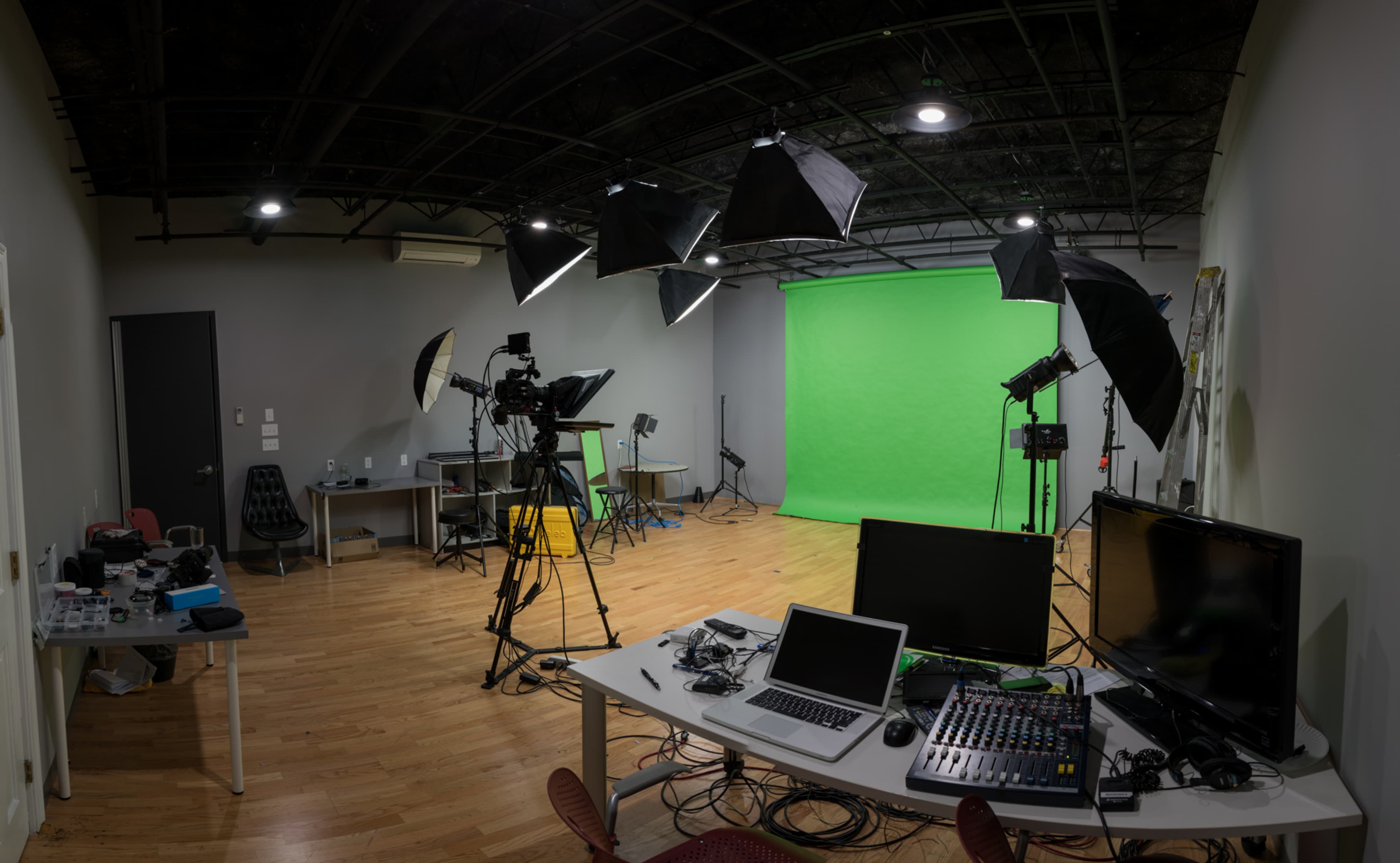 A studio setup featuring a green screen, multiple lights on stands, cameras, and a desk with computer equipment and sound mixing gear.
