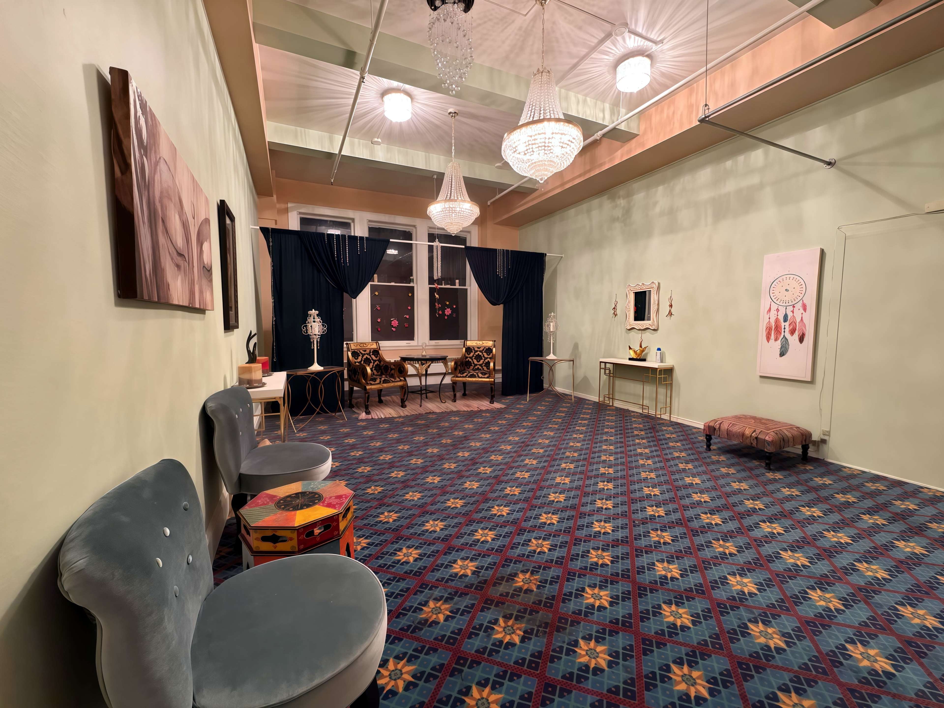 The image shows a spacious room with a patterned carpet, two decorative chairs, a table, and wall art, all illuminated by hanging chandeliers.