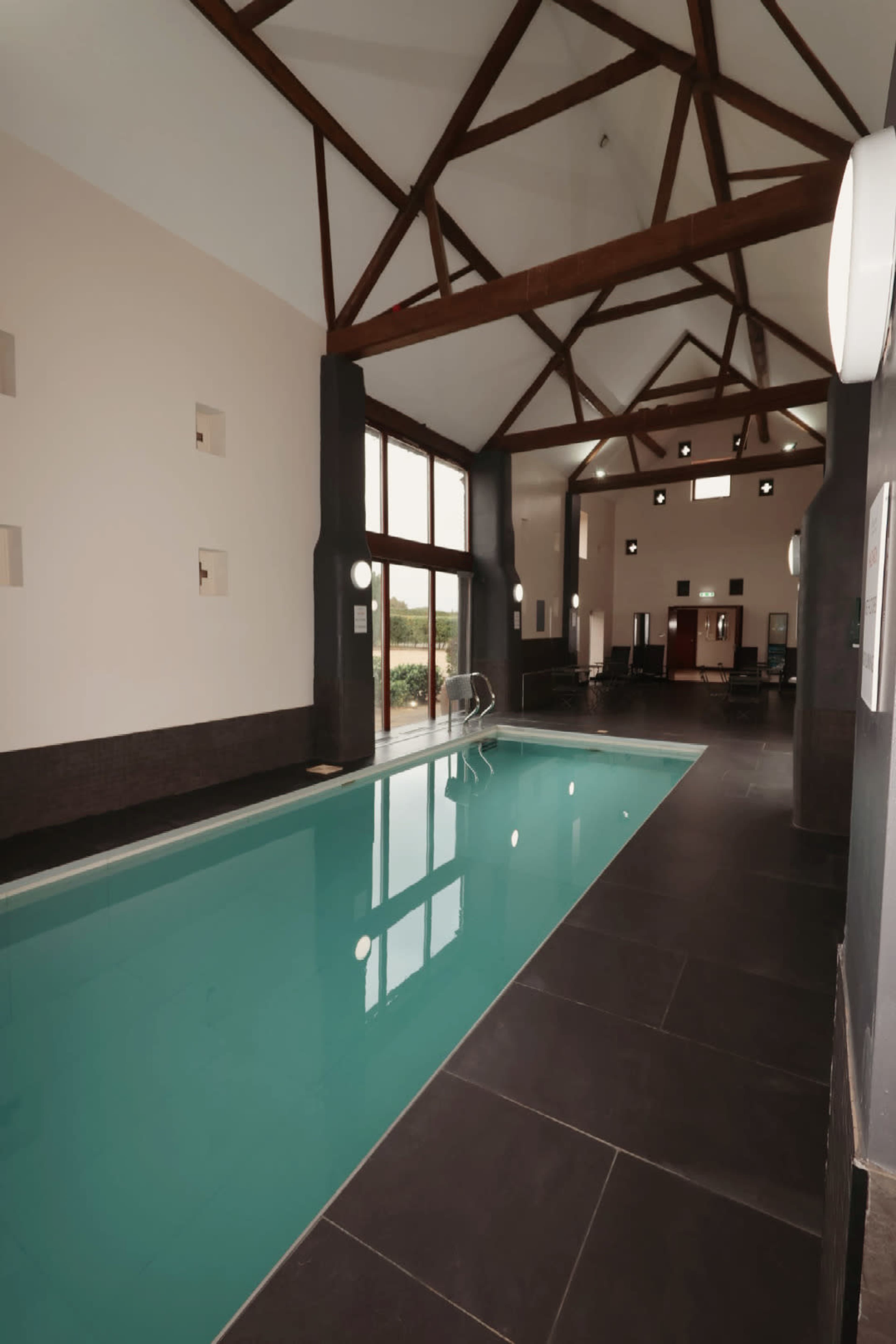 The image shows an indoor swimming pool with a turquoise surface, surrounded by large windows and a wooden beam ceiling.